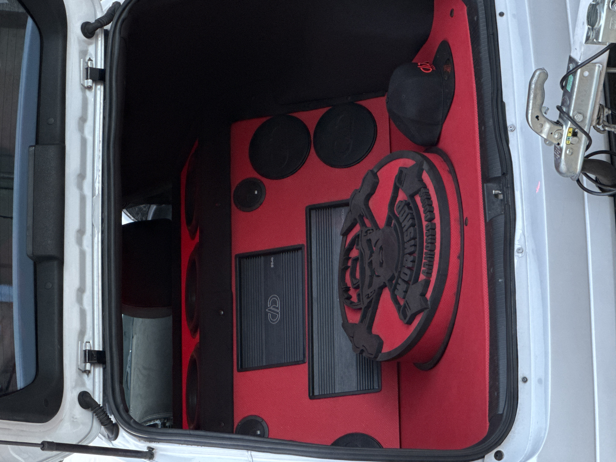 An open car trunk reveals a custom sound system installation. The interior is lined with red fabric, featuring multiple speakers and two audio amplifiers mounted on the back panel. A circular, black and red emblem with a cartoonish skull design is positioned on the floor of the trunk. A black baseball cap with a red logo rests next to the emblem. The vehicle appears to be a white hatchback, and the image is taken from behind, looking into the trunk. In the foreground, a silver trailer hitch assembly is visible. The scene is consistent with a car parked in an outdoor setting, potentially a residential area or a car show, given the customization. The specific location in Busmantsi, Bulgaria, is identified by context not visible in the image itself.