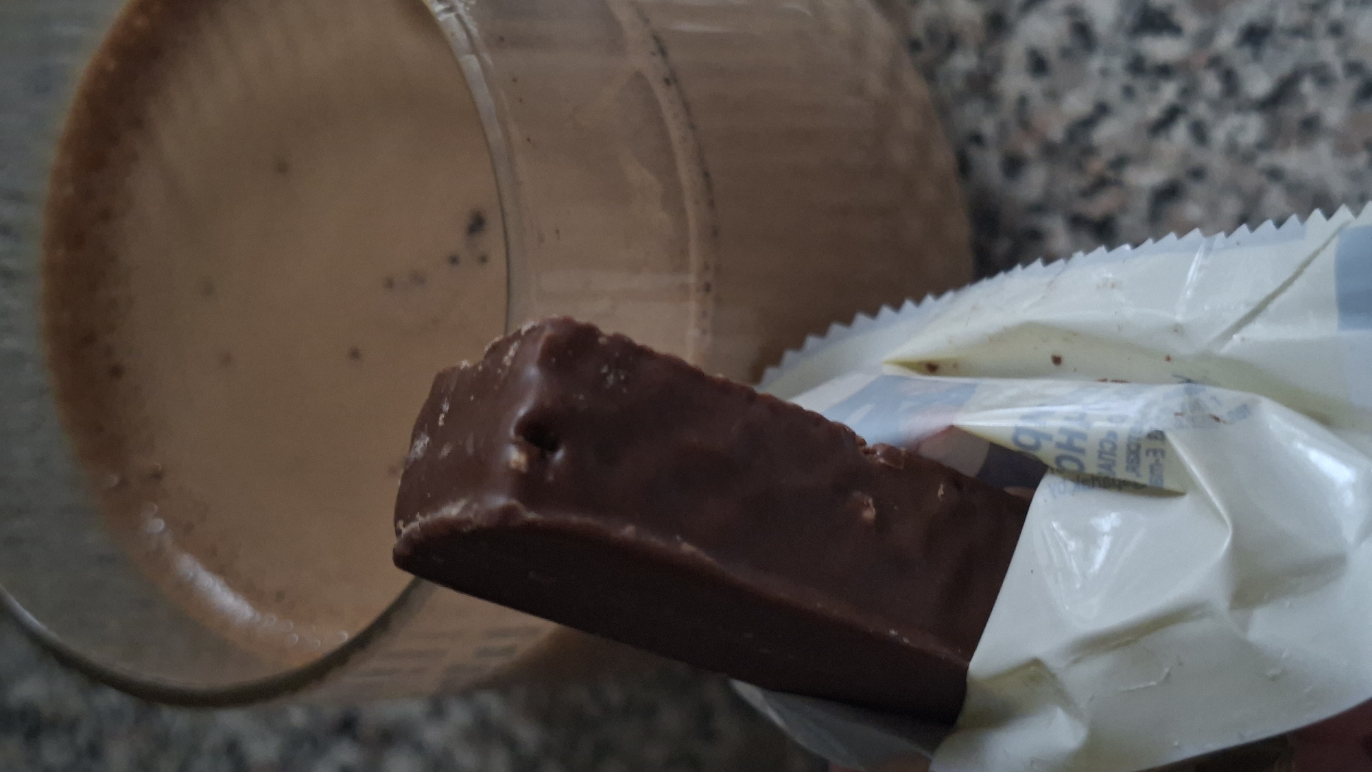 A chocolate bar is held in front of a glass of chocolate milk. The chocolate bar is partially unwrapped, revealing its brown, foil-like packaging. The chocolate bar itself is dark brown with some unevenness on its surface. The glass of chocolate milk is in the background, filled to near the top, with a frothy layer on its surface and some small bubbles. The scene appears to be set on a speckled countertop. The image does not provide any information about the city of Myadzyel, Belarus.