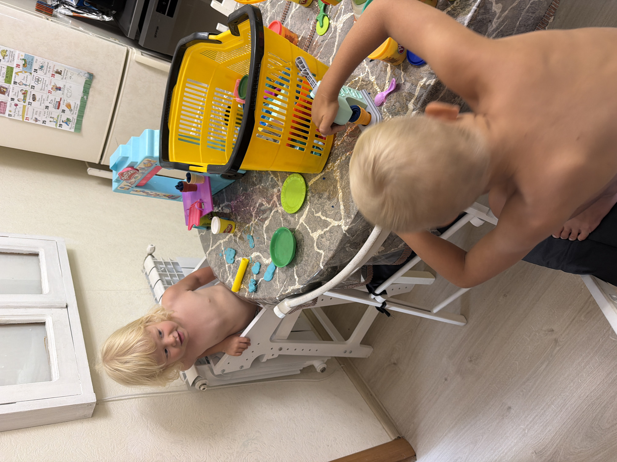 In this image, two young children, a boy and a girl, are engaged in playful activities at a table. The girl, with blonde hair, is seated in a high chair, looking directly at the camera with a smile. The boy, also with blonde hair, is standing and leaning over the table, interacting with toys.
The table is covered with a marble-patterned tablecloth and is strewn with various colorful toys, including a bright yellow shopping basket, Play-Doh containers, and other plastic play items. A small blue Play-Doh playset is also visible. The children appear to be enjoying their playtime, with the overall atmosphere seeming relaxed and cheerful.
In the background, a white refrigerator is visible with a calendar attached to it, showing dates from 17 to 30. Some text in Russian is visible on the calendar, including dates and what appear to be event descriptions. A kitchen appliance, possibly a stove, is also partially visible above the refrigerator. The setting appears to be a kitchen or dining area within a home. The lighting suggests it is daytime. The image captures a candid moment of childhood play and interaction.