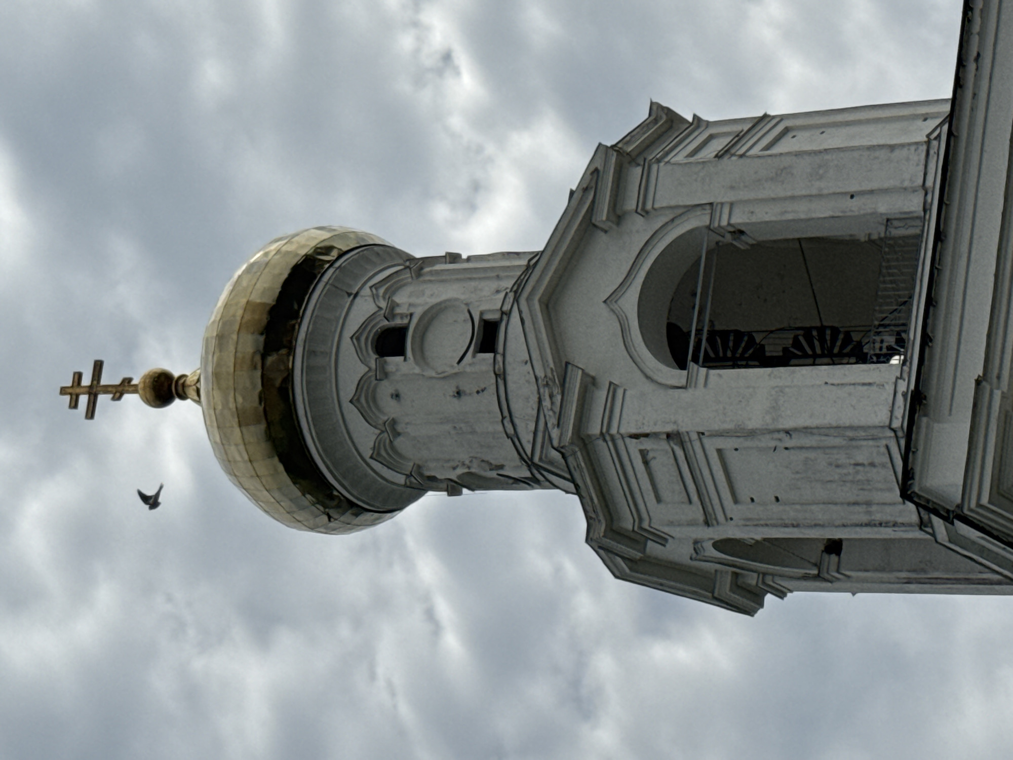 The image shows a low-angle shot of a bell tower in Veliky Novgorod, Russia. The bell tower is made of white stone and has a golden dome topped with a cross. A bird is flying in the cloudy sky above the tower. The bell tower has arched openings and architectural details typical of Russian Orthodox churches. The spiral staircase inside one of the openings is visible.
