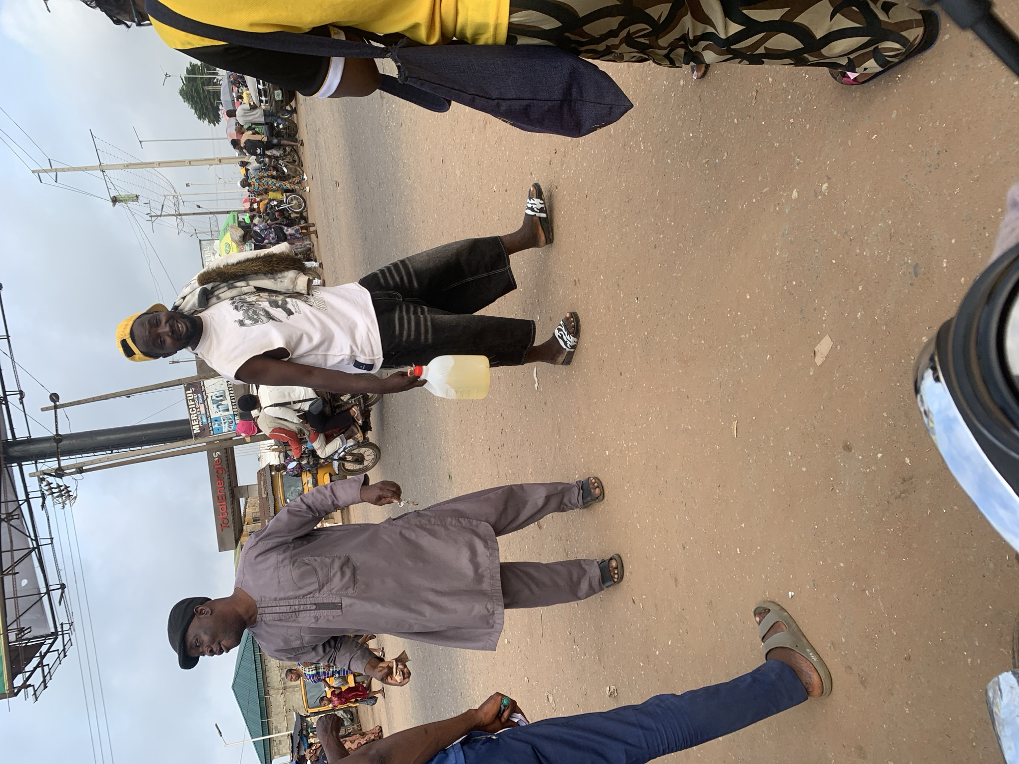 The image captures a street scene in Shagamu, Nigeria, likely near a TotalEnergies gas station. Two men are prominent in the foreground. The man on the left wears a grey traditional outfit and a black cap, and appears to be speaking or gesturing. He is holding a small wad of cash. To his right, a man in a white sleeveless t-shirt, black shorts, and a yellow cap holds a clear plastic jug filled with a light yellow liquid, possibly fuel or water. He has a fur-lined jacket slung over his shoulder and is smiling. Behind them, various people, motorcycles, and a yellow tricycle are visible, suggesting a busy market or transportation hub. The ground is dusty and unpaved. The overall atmosphere seems to be of everyday commerce and transit in an urban African setting.