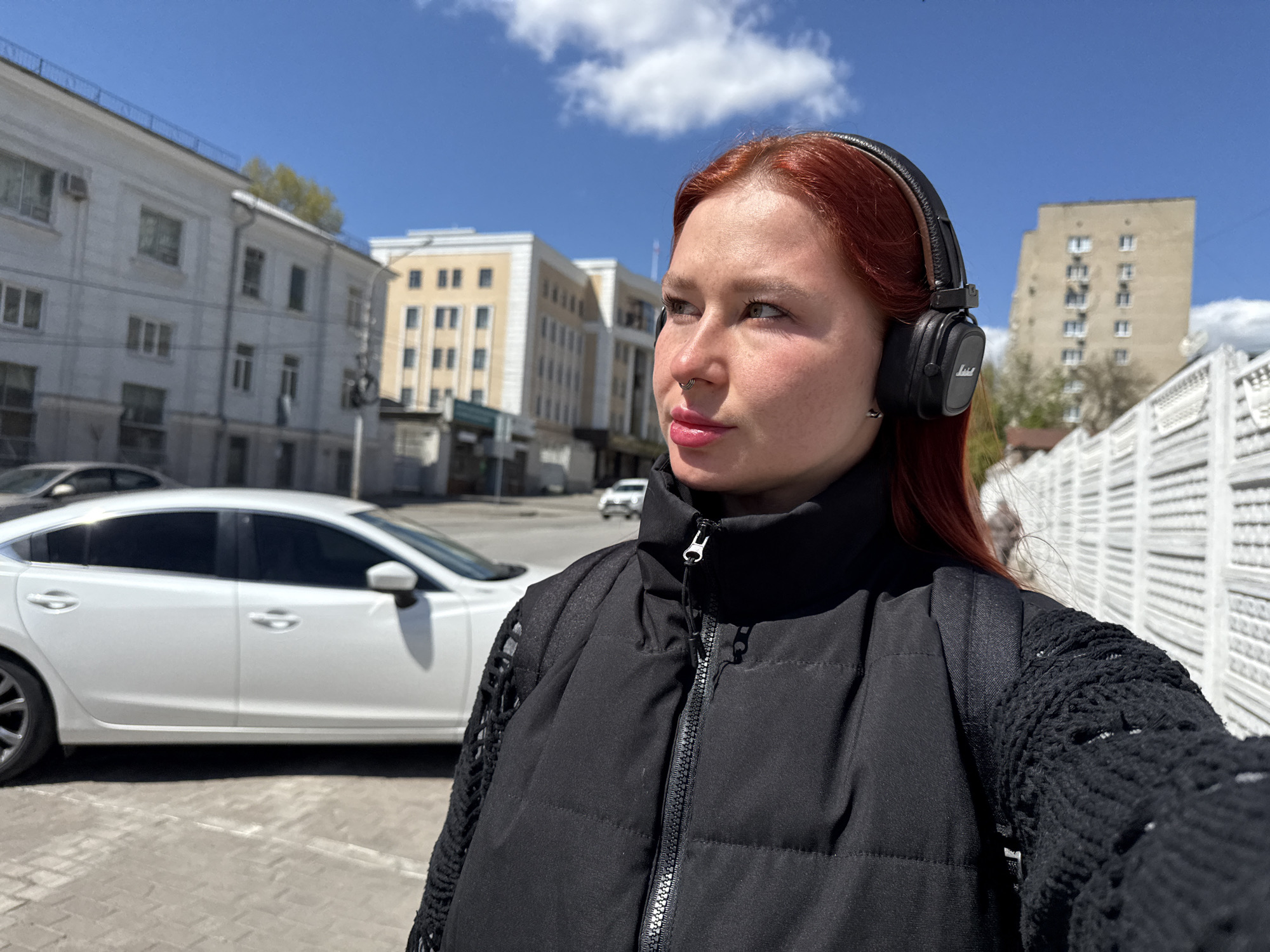 A medium shot of a woman with red hair wearing black Marshall headphones and a black puffer vest over a black crocheted sleeve shirt, is taken outdoors on a paved surface. The background reveals a city landscape under a blue sky with scattered clouds. In the immediate background to the left, a white sedan is parked. Further back are buildings, including a white structure with repeating windows and another larger beige building with multiple rows of windows. The scene is likely in Rostov-on-Don, Russia, as suggested by the architectural style of the buildings. The woman is looking slightly off-camera towards her left.