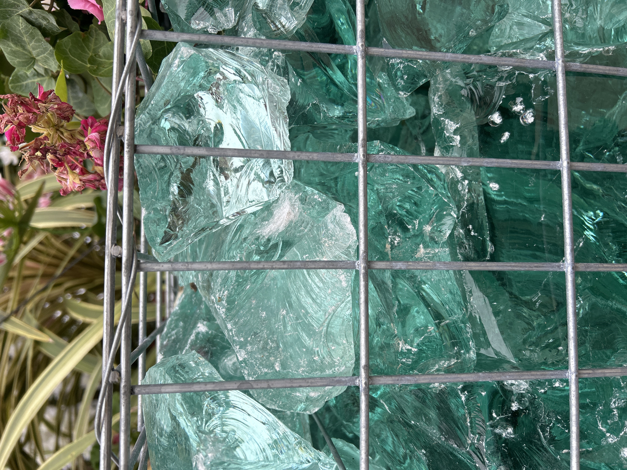 The image shows a close-up of turquoise glass pieces contained within a wire cage. A section of floral arrangement with pink flowers and green foliage is visible on the left side of the frame, partially obscured by the cage. The location in the city of Unknown, Unknown is not apparent from the image.