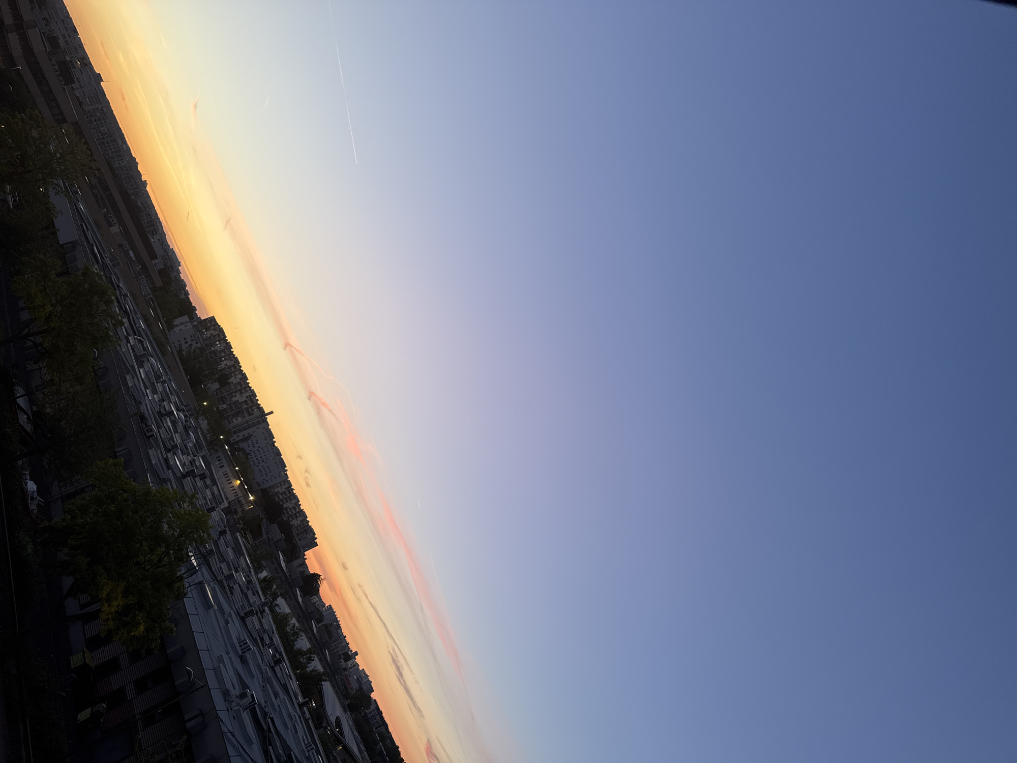 This is a landscape shot of a city skyline during sunset. The sky is a gradient of blue and orange, with thin pink clouds streaking across the horizon. Several buildings of varying heights are visible, some appearing residential and others commercial. A few trees are scattered throughout the cityscape, their leaves appearing dark against the fading light. The overall scene suggests an urban environment at dusk. The location is Unknown, Unknown.