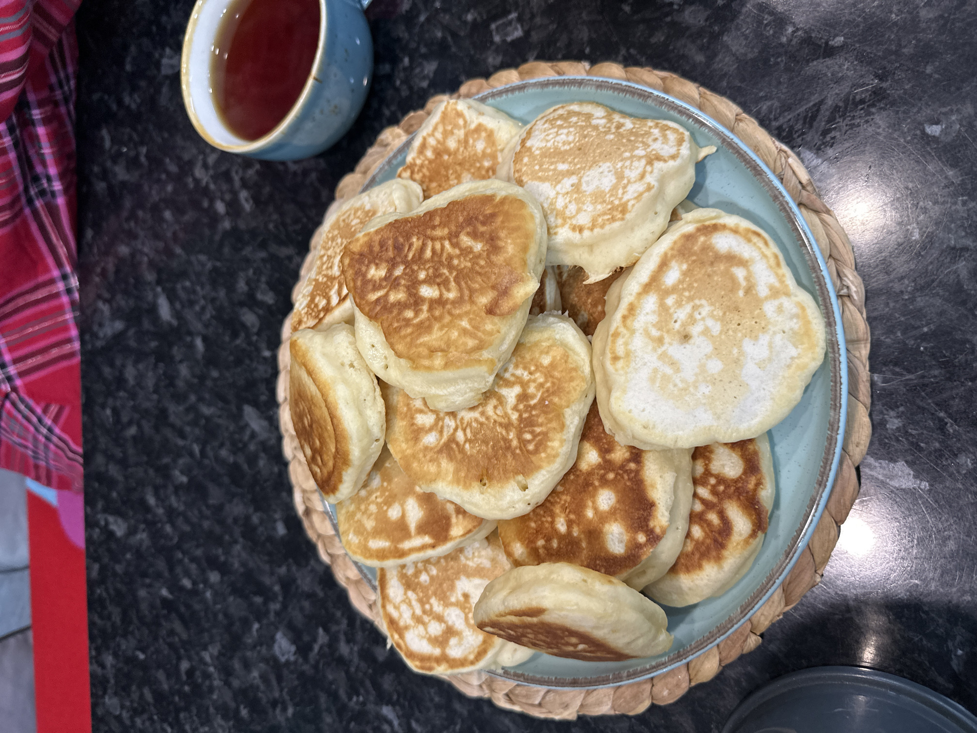 A pile of golden-brown pancakes sits on a light blue plate, which rests on a woven placemat. A light blue mug filled with a dark red liquid, likely tea, is positioned to the right of the plate. The background consists of a dark, speckled countertop. Above the countertop, a red and pink plaid fabric is visible. There are no people or animals in the image. The scene appears to be indoors, on a kitchen counter. The image does not contain any information about the city of Sochi, Russia.