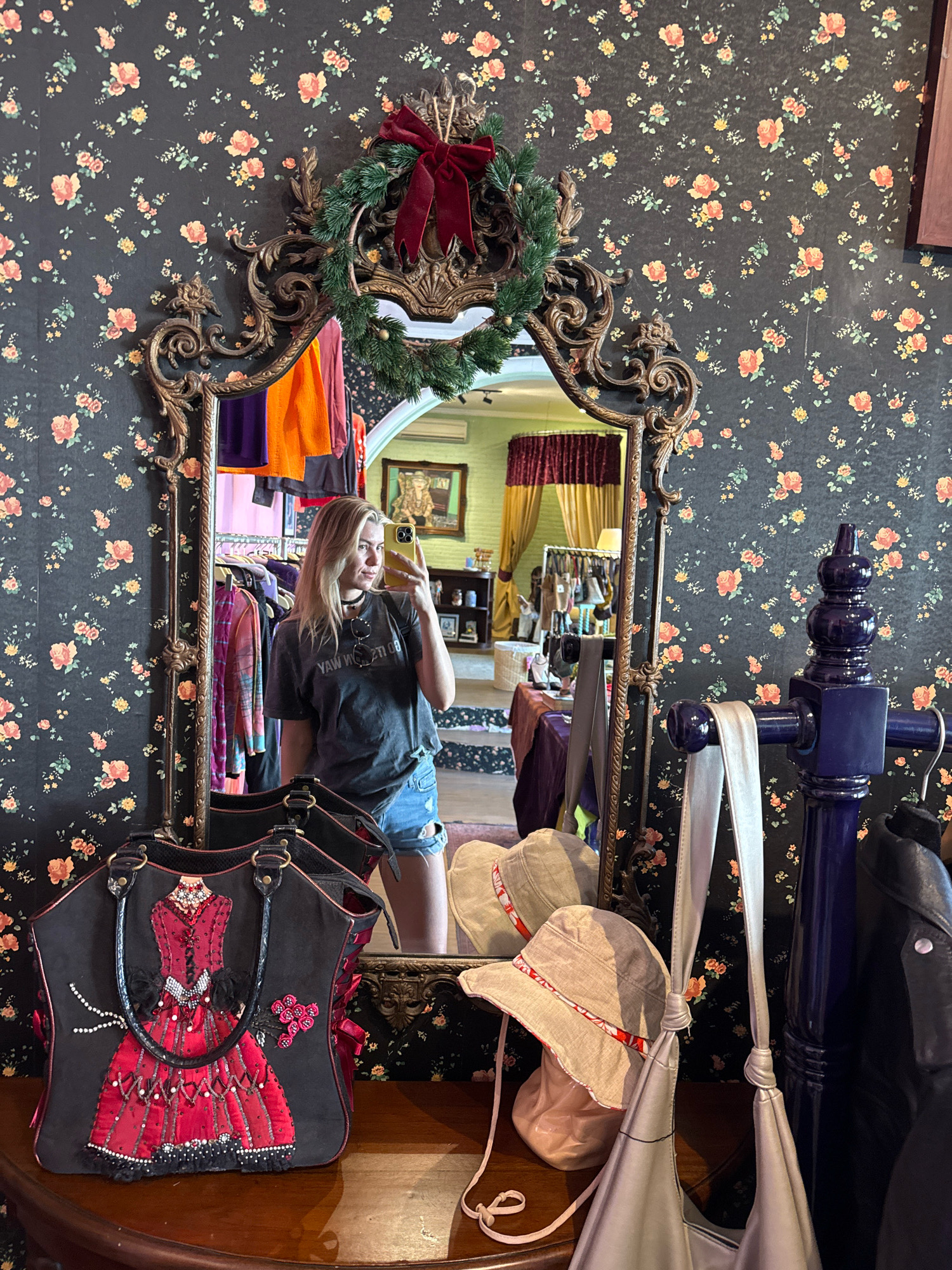 A young woman with blonde hair takes a mirror selfie in what appears to be a boutique. She is wearing a dark grey t-shirt and denim shorts. She has a choker necklace and sunglasses on her head. Behind her, the wall is covered in dark floral wallpaper. A large, ornate mirror with a Christmas wreath and bow is in the center of the frame. The mirror reflects clothing racks filled with apparel. In the foreground, there are several handbags and hats displayed on a wooden surface. To the right, a tall, dark blue post is visible with a black leather jacket hanging on it. The overall impression is of a retail environment with a touch of holiday decor.