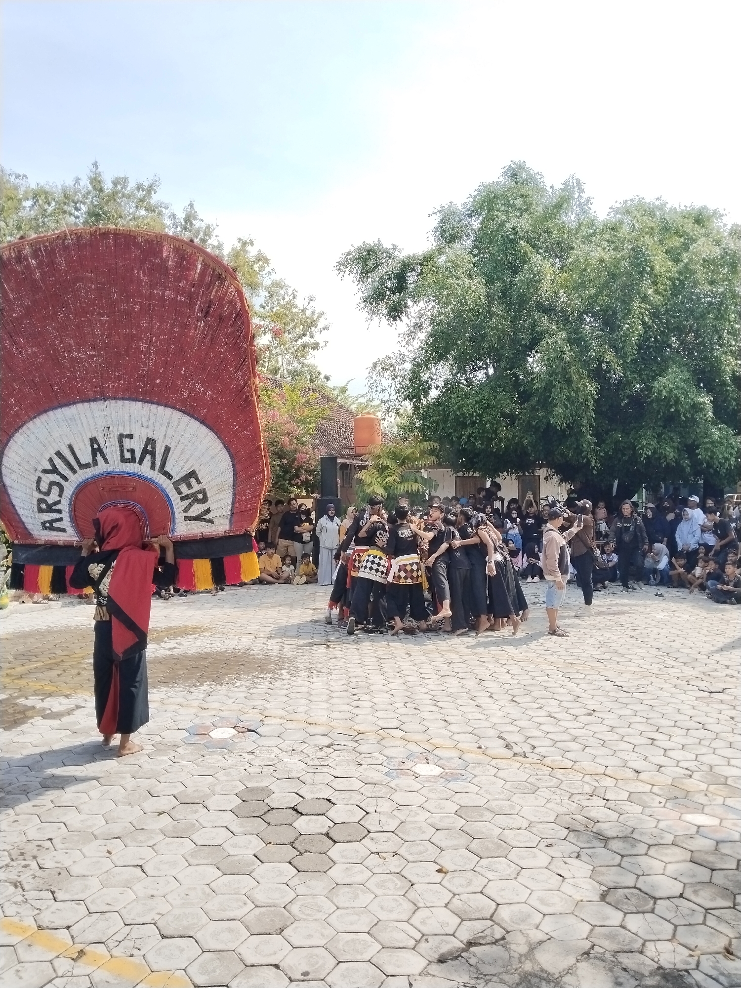 The image captures a cultural performance in Karangmalang, Indonesia. A person stands in the foreground, carrying a large, circular banner with the words "ARSXYILA GALERY" printed on it. A group of performers dressed in black attire is gathered in the center of the scene. A crowd of spectators surrounds the performers, with some people sitting and others standing. The performance takes place on a paved courtyard, with buildings and trees in the background. The sky is bright and clear.