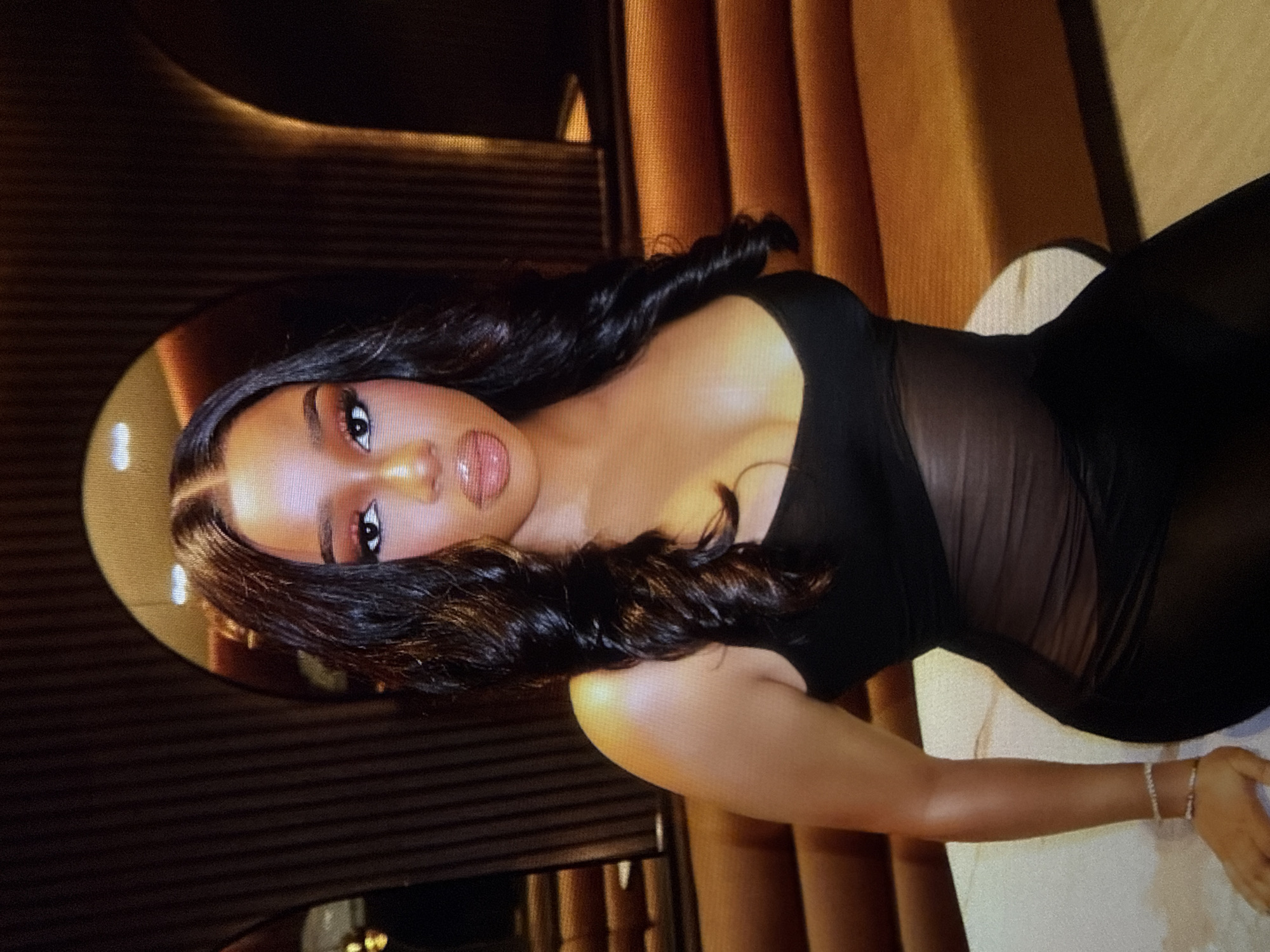 A medium shot shows a Black woman posing for a photo indoors. She has long, dark, wavy hair and is wearing full makeup, including eyeshadow, eyeliner, mascara, blush, and lip gloss. Her dress is black and appears to be sheer from the mid-torso down. A portion of her right arm is visible, adorned with a bracelet on her wrist and another on her ankle. Her left leg is bent at the knee, with her foot resting on a white surface. The background features a dark, textured wall with arched openings, and orange upholstered seating. The lighting suggests an evening setting, possibly a restaurant or lounge. This scene is not located in Abuja, Nigeria, as there are no visual cues to support this claim.