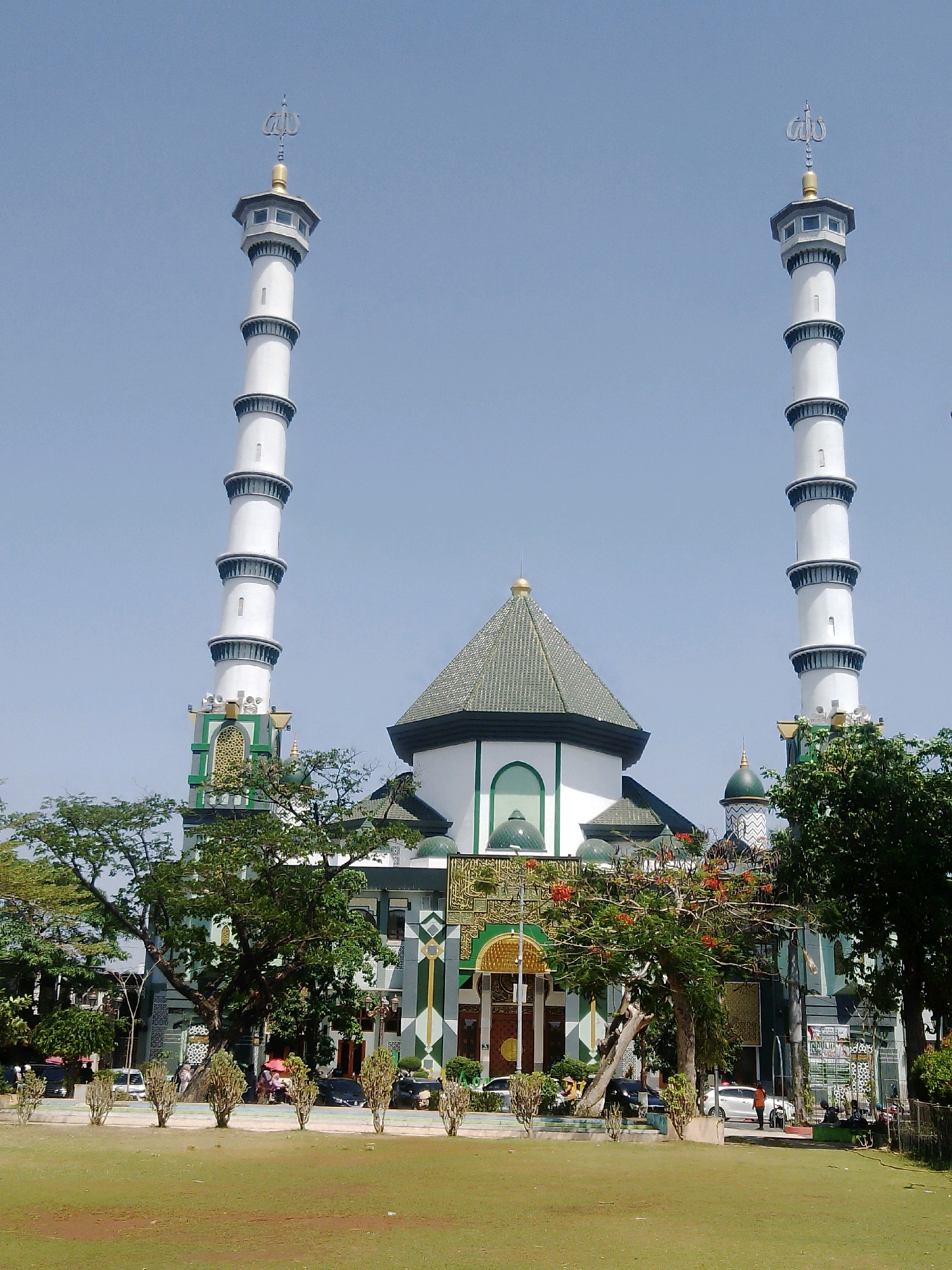 The image shows a front view of the Islamic Center of Lamongan, located in Lamongan, East Java, Indonesia. The building features two prominent white minarets with multiple tiers and golden finials topped with Arabic calligraphy. A central dome structure with a green roof and a smaller green dome are also visible. The facade of the mosque is adorned with green and gold geometric patterns and Arabic script. In front of the mosque, there is a well-maintained green lawn and a paved area with several trees, including one with red flowers. A few cars are parked in the background, and several people are seen standing or sitting near the entrance and on the side. The sky is clear and blue, indicating a sunny day.