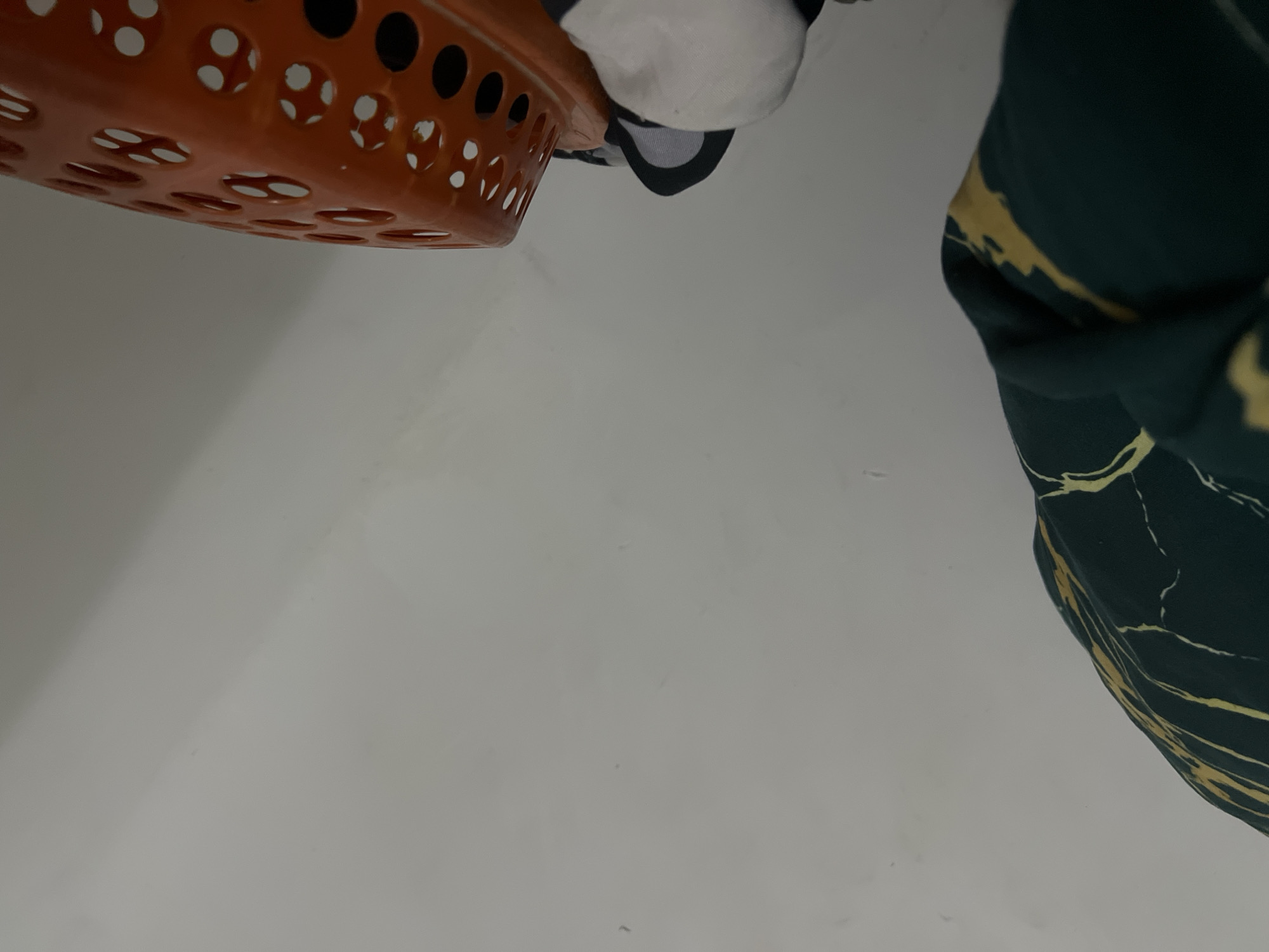 The image shows a close-up of a person's legs and feet, wearing socks and standing next to an orange plastic laundry basket. A dark green garment with yellow marbling is draped over the left side of the frame. The background is a plain, light-colored wall. The scene appears to be indoors, possibly in a bedroom or laundry area. There is no information in the image to indicate a specific location in the city of Howayi, Nigeria.