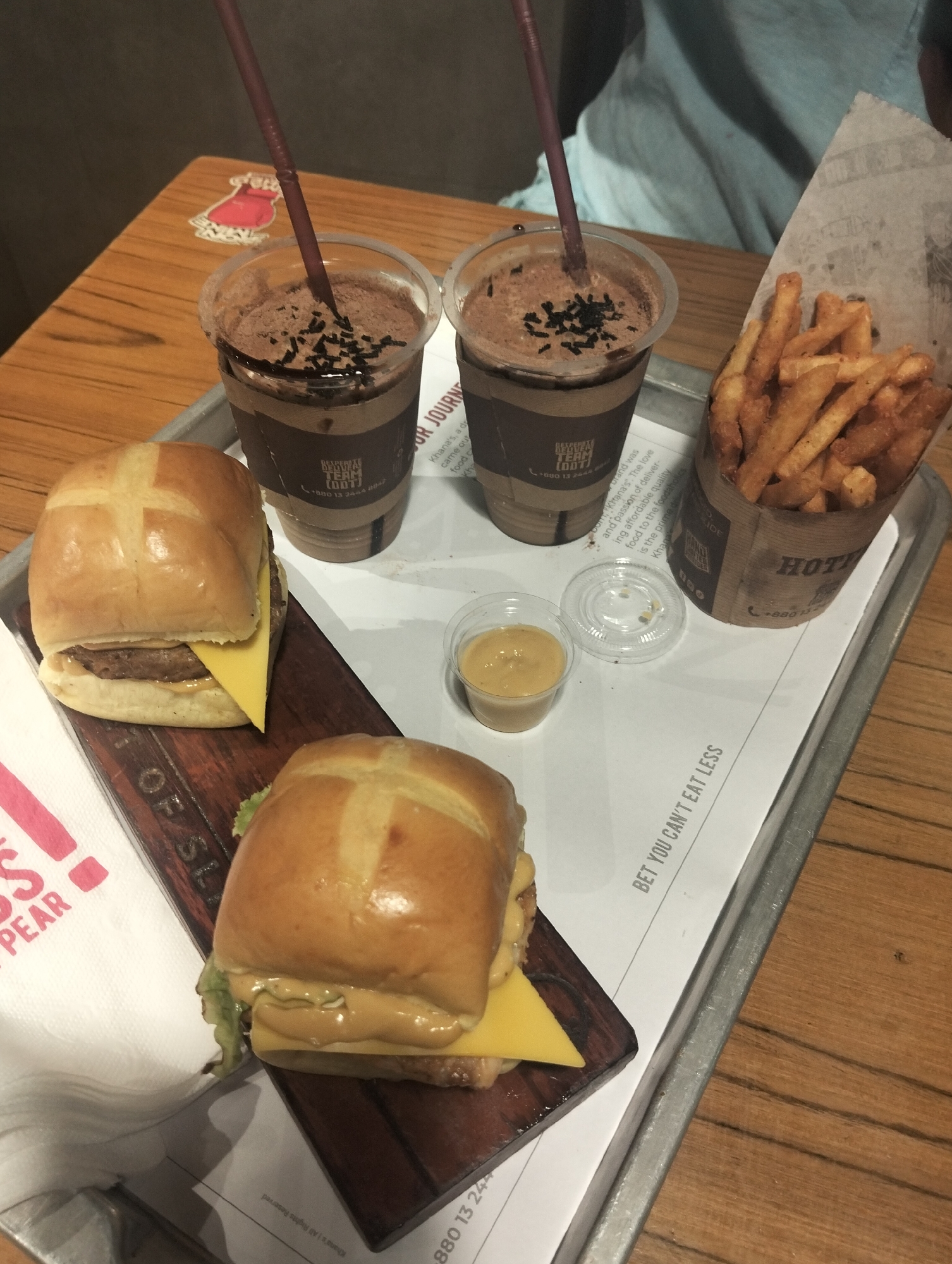A high-angle shot captures a tray laden with fast food, likely in Dhaka, Bangladesh, considering the phone number prefixes visible (+880). Two cross-bun topped burgers, each with a slice of yellow cheese, are presented on a dark wooden board. The tray also holds two chocolate milkshakes with dark sprinkles, served in transparent plastic cups with brown sleeves and accompanied by straws. A container of French fries sits upright in a paper cup next to a small, transparent plastic container filled with a yellow condiment. The tray is lined with white paper printed with text in black. The table is made of wood, and a person wearing a light blue top is partially visible in the background. A red sticker is on the left of the milkshake.