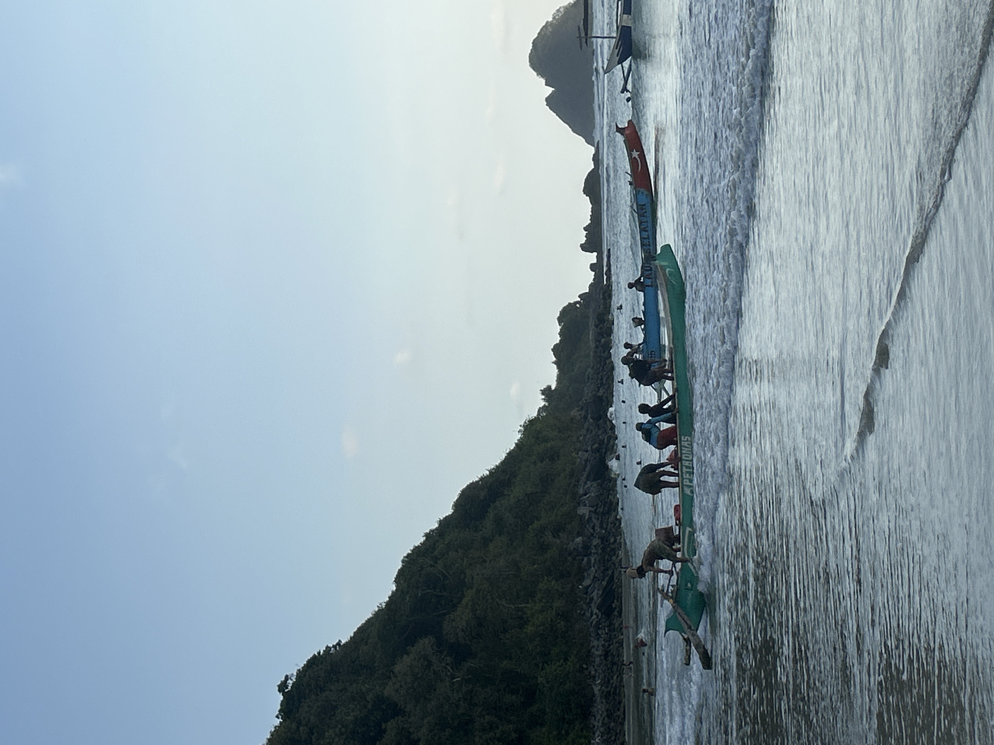 A group of people are in and around several traditional fishing boats in shallow ocean water near a beach. In the foreground, a green boat with the word "PETRONAS" visible on its side is being pushed or pulled by individuals. Further back, a red and blue boat with text that appears to read "LIHU SELATAN" is also in the water with people aboard. Several other boats are visible further out in the water, and some people are swimming or wading near the shore. Behind the beach, a lush green, tree-covered hillside slopes down towards the water. Rocky outcrops and larger islands are visible in the distance. The sky is clear and pale blue. The scene is set at Selongbelanak Beach, Lombok, Indonesia.