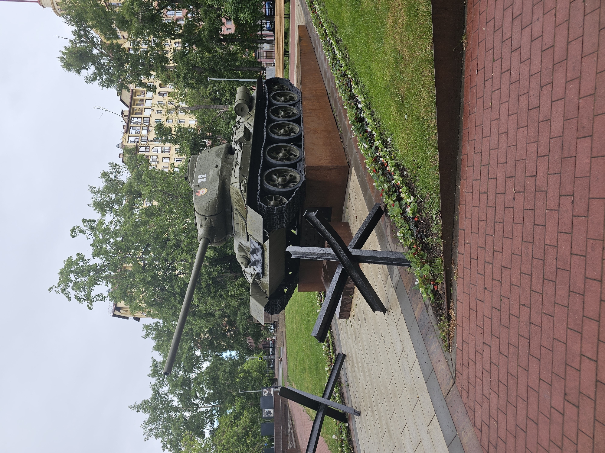 The image shows a T-34 tank monument located in Khimki, Russia. The tank, painted in green, is positioned on a concrete base, with steel anti-tank obstacles placed in front of it. The number "22" and a red star are visible on the turret. Behind the tank, there's a grassy area with trees and a multistory building in the background. The foreground includes a brick walkway and a narrow strip of land with grass and flowers.