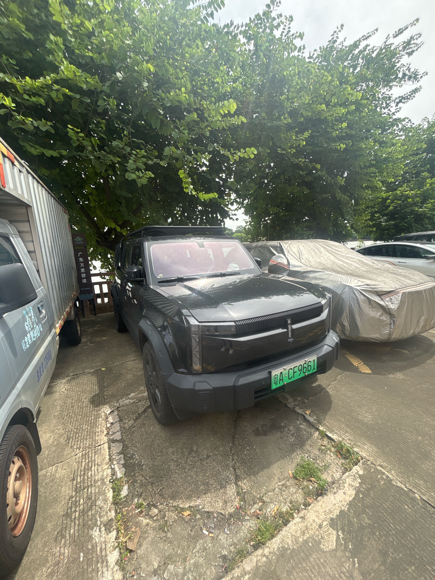 In Nanhai District, China, an outdoor scene captures a dark-colored SUV parked on a concrete surface. The SUV's license plate reads "A-CF9661". To the left of the SUV is a portion of a silver truck. Behind the SUV, a car is covered with a light-colored tarp. Lush green trees with dappled sunlight overhead partially shade the vehicles. The ground shows cracks and some patches of grass growing through.