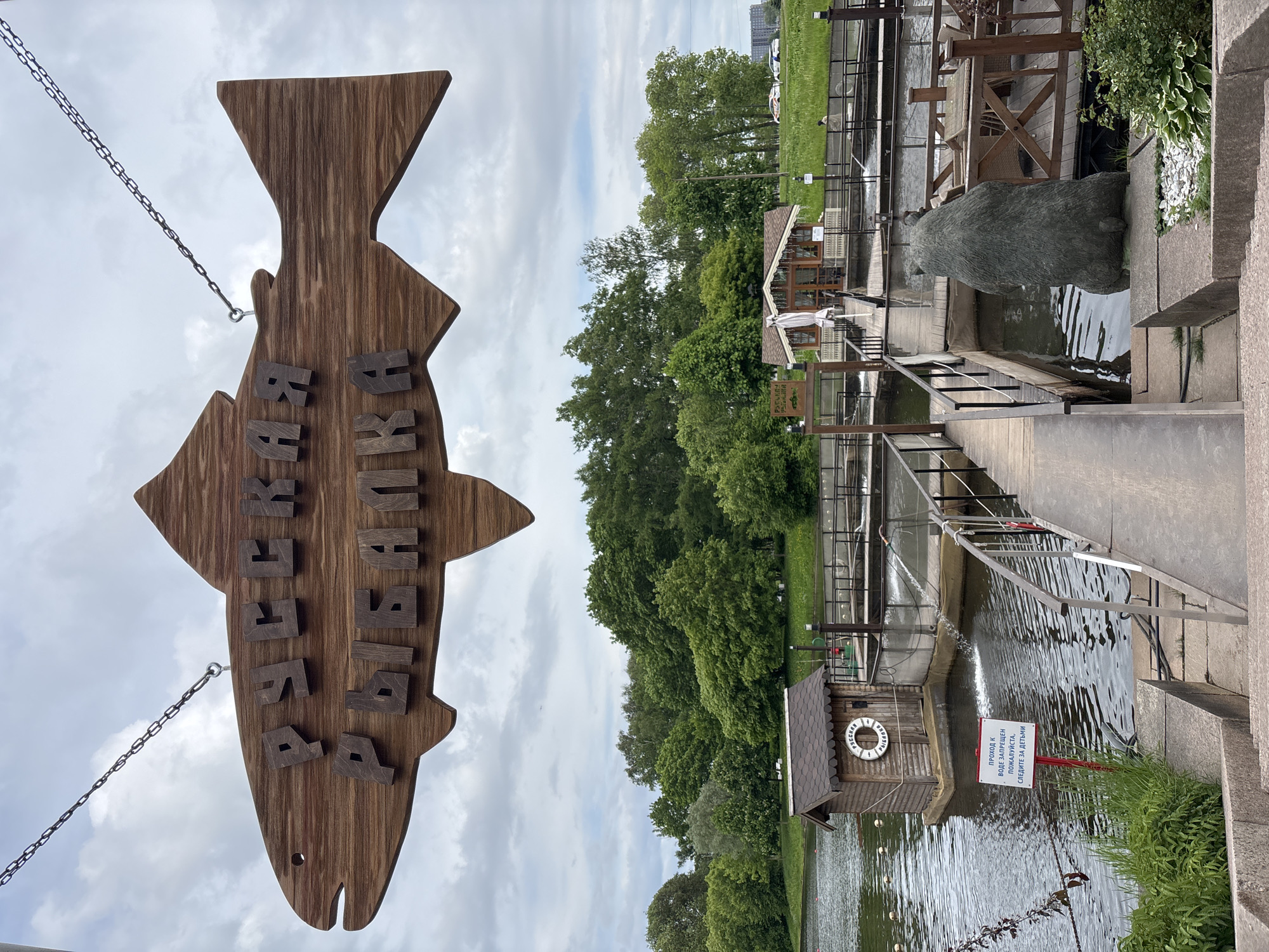 This outdoor image captures a recreational area with a strong fishing theme under a cloudy sky. In the left foreground, a large, dark wooden sign shaped like a fish hangs by chains. The sign bears raised wooden letters in Russian, reading "РЫБАЛКА И РЫБКА," which translates to "FISHING AND FISH" or "FISHING AND SMALL FISH."
The midground and background on the right side of the image feature a complex of interconnected water channels or ponds, flanked by wooden walkways and bridges. Several small wooden structures with sloped roofs are visible, one of which has a lifebuoy attached, inscribed with "СПАСАТЕЛЬНЫЙ КРУГ" (LIFEBUOY). Near the water, a prominent dark statue of a bear is positioned, seemingly looking into the water. Further along the water's edge, a red sign warns in Russian: "ПРОХОД К ВОДЕ ЗАПРЕЩЕН, ПОЖАЛУЙСТА, СЛЕДИТЕ ЗА ДЕТЬМИ!" (ACCESS TO WATER IS FORBIDDEN, PLEASE, WATCH YOUR CHILDREN!).
Beyond the walkways and water, lush green trees dominate the background, suggesting a park or forested area. Faintly visible in the far distance, above the trees, are a few cars on what appears to be a road or parking lot. Outdoor seating areas with tables and chairs under awnings can be seen on some of the platforms, hinting at a cafe or restaurant. The setting appears peaceful and designed for leisure, likely a fishing park or a riverside establishment in Saint Petersburg, Russia, given the context and visible text. The time of day is daytime, with overcast weather indicated by the uniformly cloudy sky.