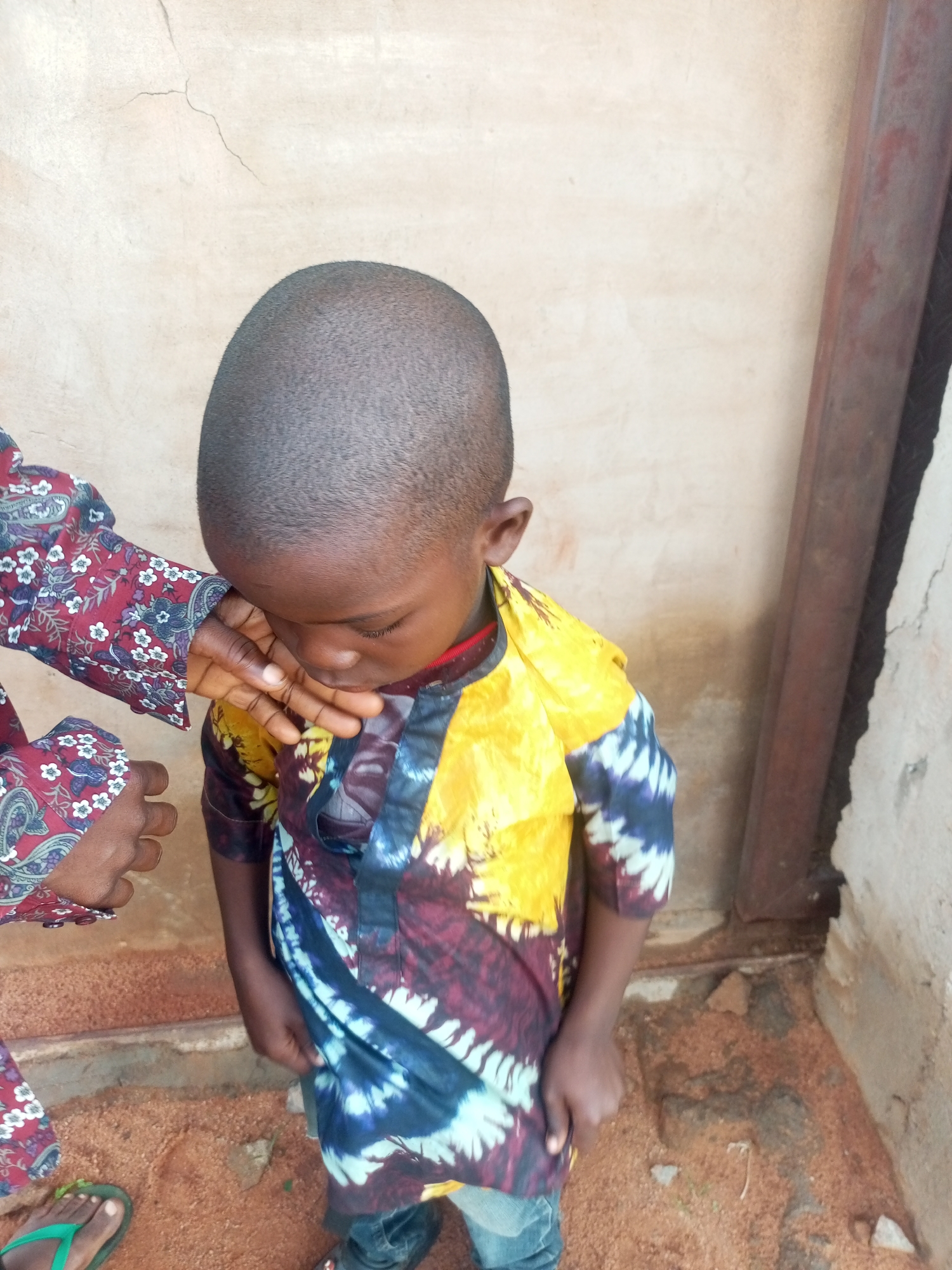 A young Black child with a shaved head is looking down, with their face partially obscured by a hand. The child is wearing a vibrant, multi-colored dashiki with yellow, purple, and blue tie-dye patterns. A person, likely an adult, is partially visible on the left, wearing a patterned shirt with red, white, and blue floral designs. Their hand is extended towards the child, touching their mouth or cheek. The background consists of a textured, beige wall with a crack near the top, and a dark, vertical post on the right side. The ground is covered in reddish-brown dirt and small stones. The scene appears to be outdoors, possibly in a courtyard or alleyway. The overall impression is an intimate or comforting interaction between the child and the adult. This scene is likely located in a residential area within Tempure, Nigeria.