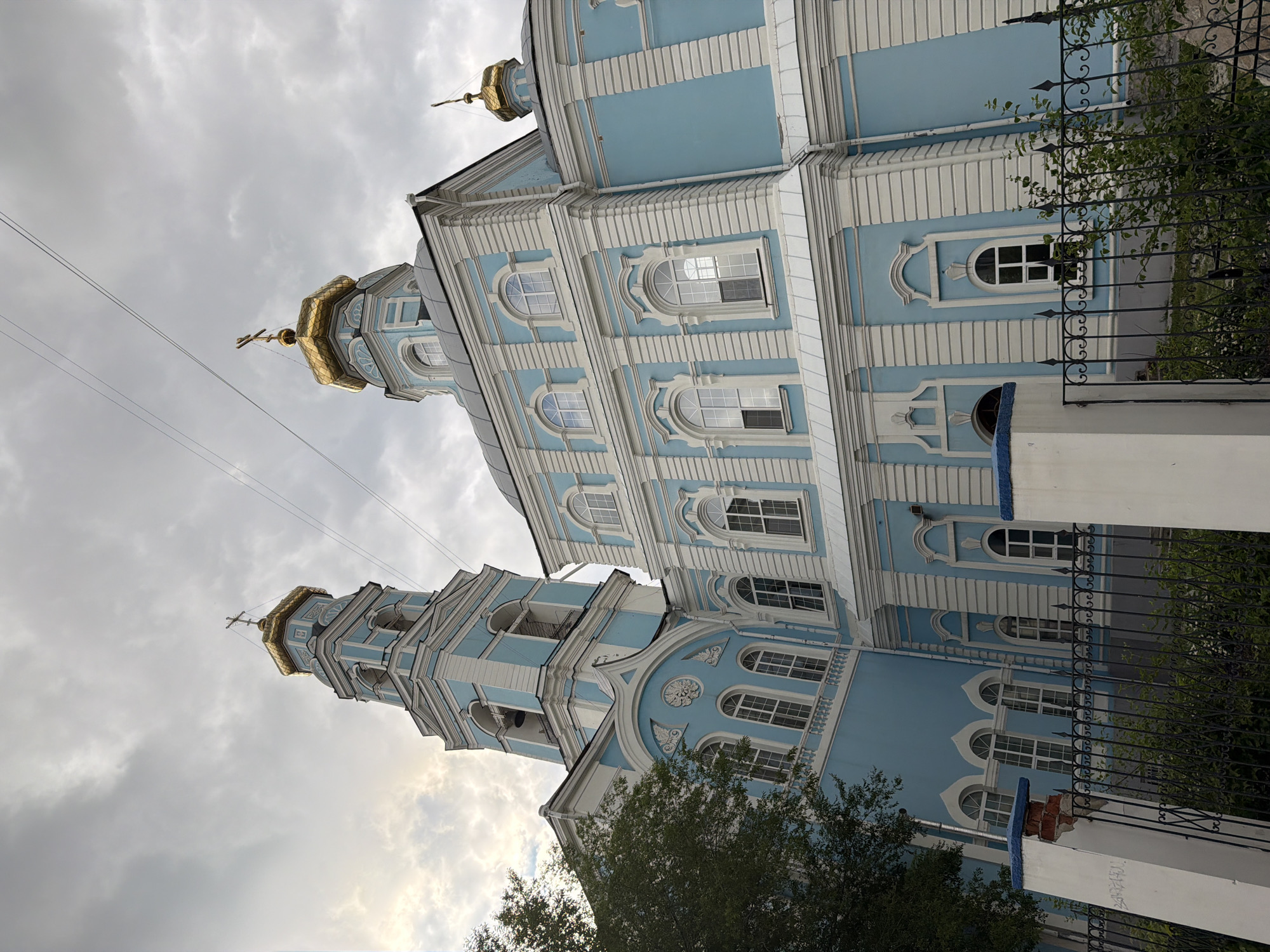 The image displays a majestic, light blue and white Orthodox church under an overcast sky. The church features prominent golden onion domes, each crowned with a golden Orthodox cross, signifying its religious purpose. The building's architecture is ornate, characterized by multiple stories adorned with white decorative elements such as pilasters, cornices, and intricately framed arched windows against the blue facade.
Two main structures are visible: a larger section on the right with a golden dome and multiple tiers of windows, and a slightly smaller bell tower on the left, also topped with a golden dome and cross. The bell tower has open arched sections, suggesting where bells would be housed.
In the foreground, a black wrought iron fence with spiked finials partially obscures the lower section of the church, and some green foliage from trees or bushes is visible, particularly near the base of the bell tower and behind the fence. Power lines are faintly visible stretching across the cloudy sky in the upper left portion of the frame. The cloudy weather suggests a subdued daytime atmosphere. No people or specific activities are depicted, focusing solely on the architectural grandeur of the structure. No text is visible in the image. Given the location context, this is an Orthodox church in Yekaterinburg, Russia.