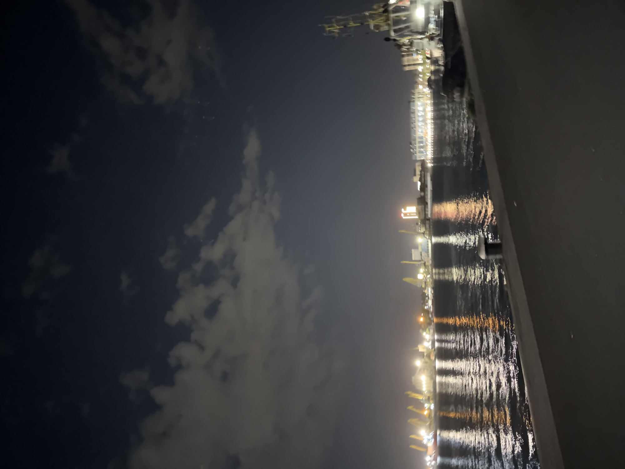 The image depicts a nighttime scene in Burgas, Bulgaria. On the right is a portion of a ship or structure, possibly a pier, with a dark grey or black exterior. To the left is a view of the Port of Burgas, visible along the water's edge. Numerous lights reflect off the water, suggesting the presence of port facilities, cranes, and buildings. The sky is dark, with scattered clouds. There are no visible people. The image appears to be taken from an elevated position on a ship.