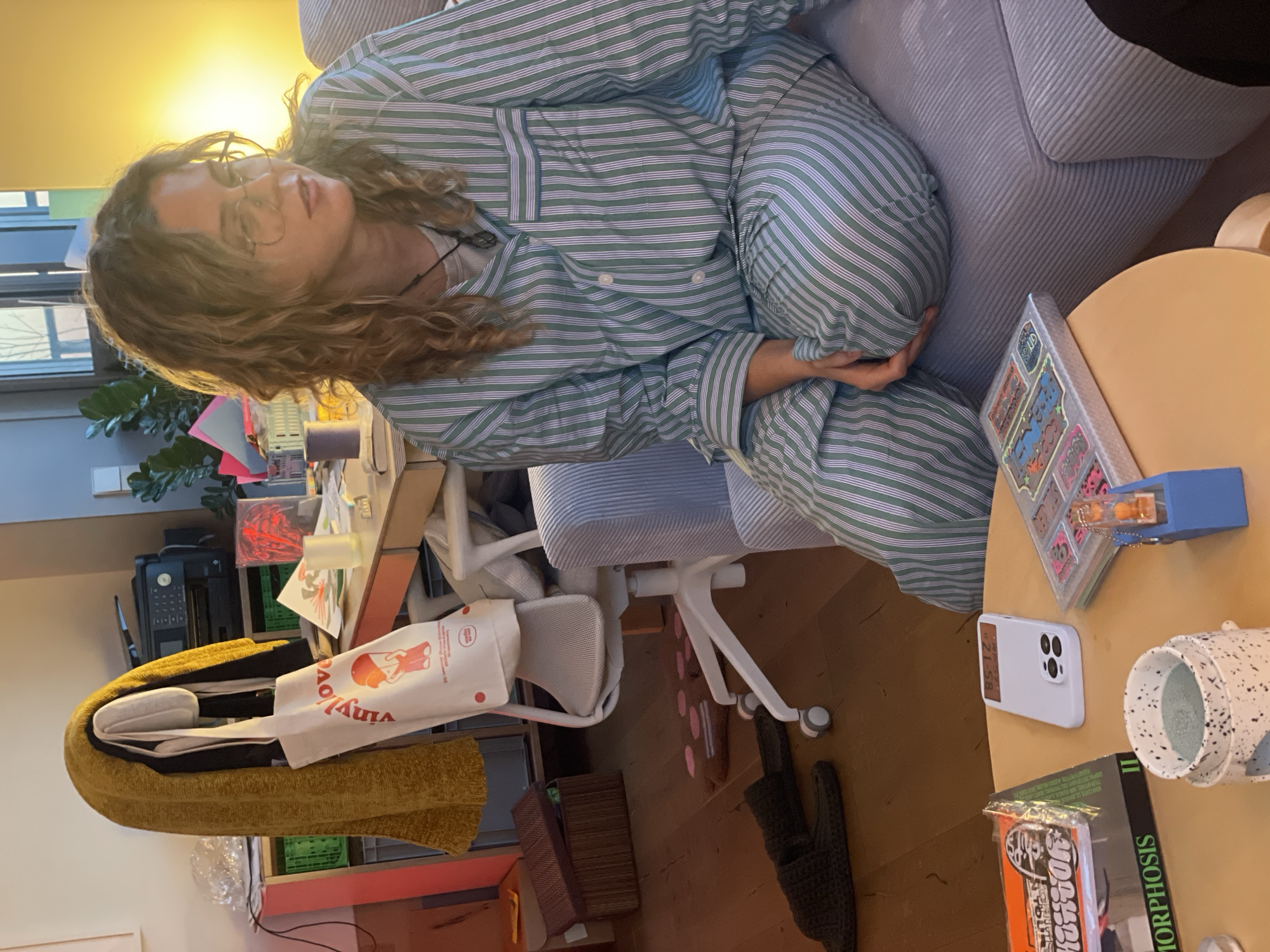 A person in striped pajamas is sitting in a grey chair with a round wooden table next to them. On the table, there is a phone, a book titled "Morphosis II," a spotted container, and a box of stickers. There is a desk and chair behind the person, along with a printer and a bag that says "vinyl love". A pair of slippers sit on the floor. The location is unknown and there is no indication this image was taken in Saint Petersburg, Russia.