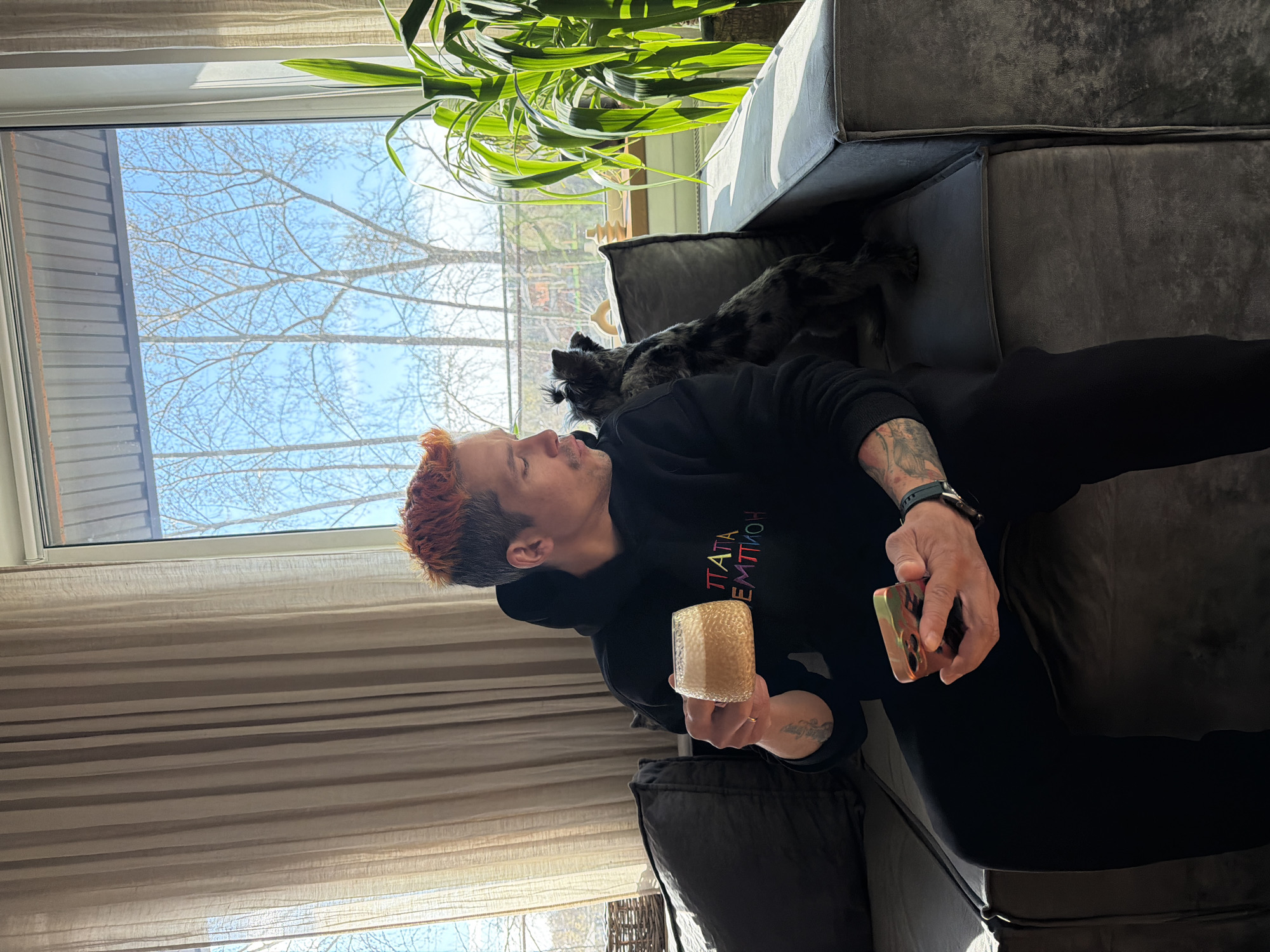 The image, taken in Chaikovsky, Russia, captures a relaxed indoor scene featuring a person and a dog on a dark-colored velvet-like couch. A male individual with orange-red hair, stubble, and visible arm tattoos is lying on his back, looking upwards and slightly to the left. He is wearing a black hooded sweatshirt, on which colorful letters spell "ПАТАТА ЕМТТИОН". In his right hand, he holds a golden, textured cup, and in his left, a smartphone with a patterned, metallic-toned case.
Resting near the person's head and chest is a small, dark-furred dog with a speckled or merle-like coat. The room is brightly lit by natural light streaming through a large window that dominates the left side of the frame. Outside, a clear blue sky is visible above bare tree branches, suggesting a late winter or early spring day. Strong sunlight casts shadows across the room, indicating it is daytime. A vibrant green houseplant is positioned in the upper right corner, and light-colored, horizontally striped curtains are drawn to the left of the window.