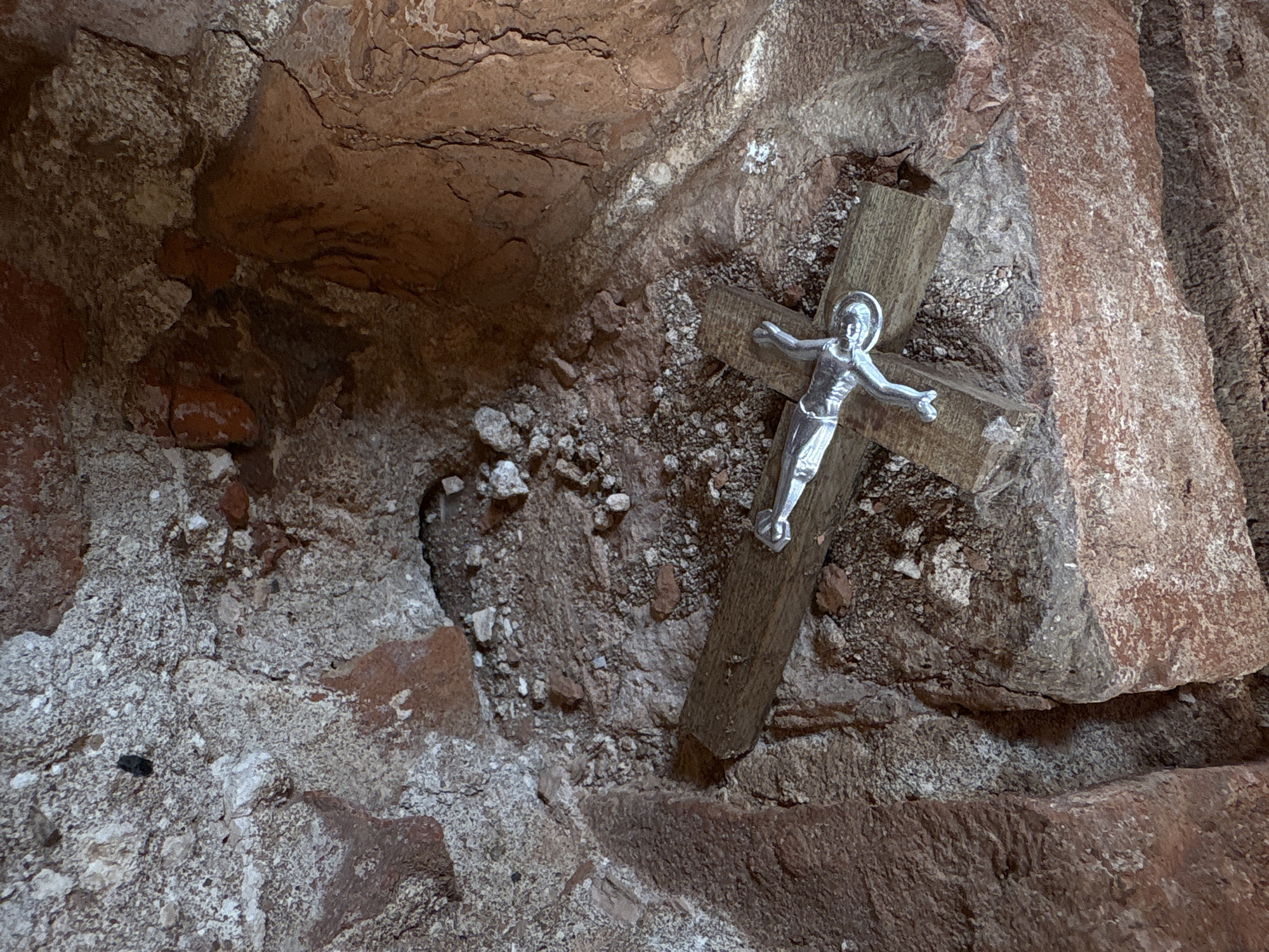 The image displays a close-up view of a crucifix nestled within a rugged, cave-like or grotto-like natural rock formation. The central object is a small crucifix, composed of a weathered wooden cross with a metallic, silver-colored corpus of Jesus Christ affixed to it. Jesus is depicted with outstretched arms, a halo around his head, and a draped loincloth.
The setting is characterized by uneven, textured rock surfaces predominantly in shades of reddish-brown, dark brown, and grayish-white. The rocks exhibit numerous cracks, crevices, and granular patches, suggesting a natural and possibly ancient environment. Loose pebbles and rock debris are visible on ledges and at the base of the crucifix, blending into the earthy surroundings.
No activity is taking place; the scene is a still life, presenting a devotional object in its environment. The lighting is diffused, suggesting an interior space or a sheltered area, rather than direct sunlight. There are no visible signs of specific time of day or weather conditions. The placement of the crucifix evokes a sense of solemnity and reverence, characteristic of a religious shrine or a personal devotional spot. The location context indicates this is in Staroe Rakomo, Russia. No visible text is present in the image.