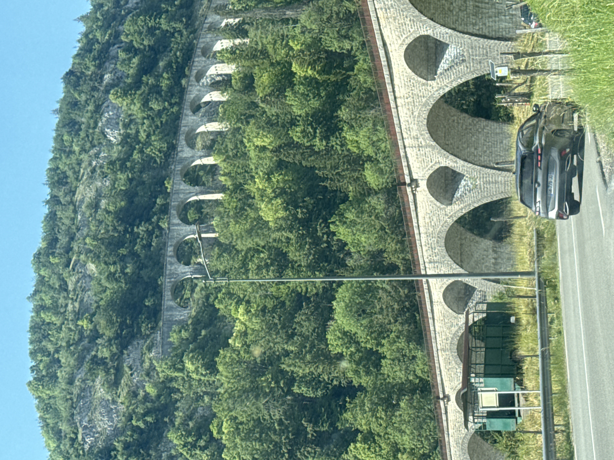 This image, taken in the vicinity of Hauts de Bienne, France, captures a scenic view of a multi-tiered stone viaduct carved into a forested mountainside under a clear blue sky. The foreground features a paved road with a dark grey SUV driving away from the viewer. To the left of the road, a green, open-sided trailer with a canopy is parked on a grassy embankment, adjacent to a metal guardrail. The prominent viaduct, constructed from light-colored stone arches, spans across the frame, with a second, higher viaduct visible further up the slope. Dense green trees cover the mountainside, with some rocky outcrops interspersed. A tall, slender lamp post stands near the lower viaduct on the right side of the road. The overall environment is rural and mountainous, characterized by extensive vegetation and historic architectural structures.