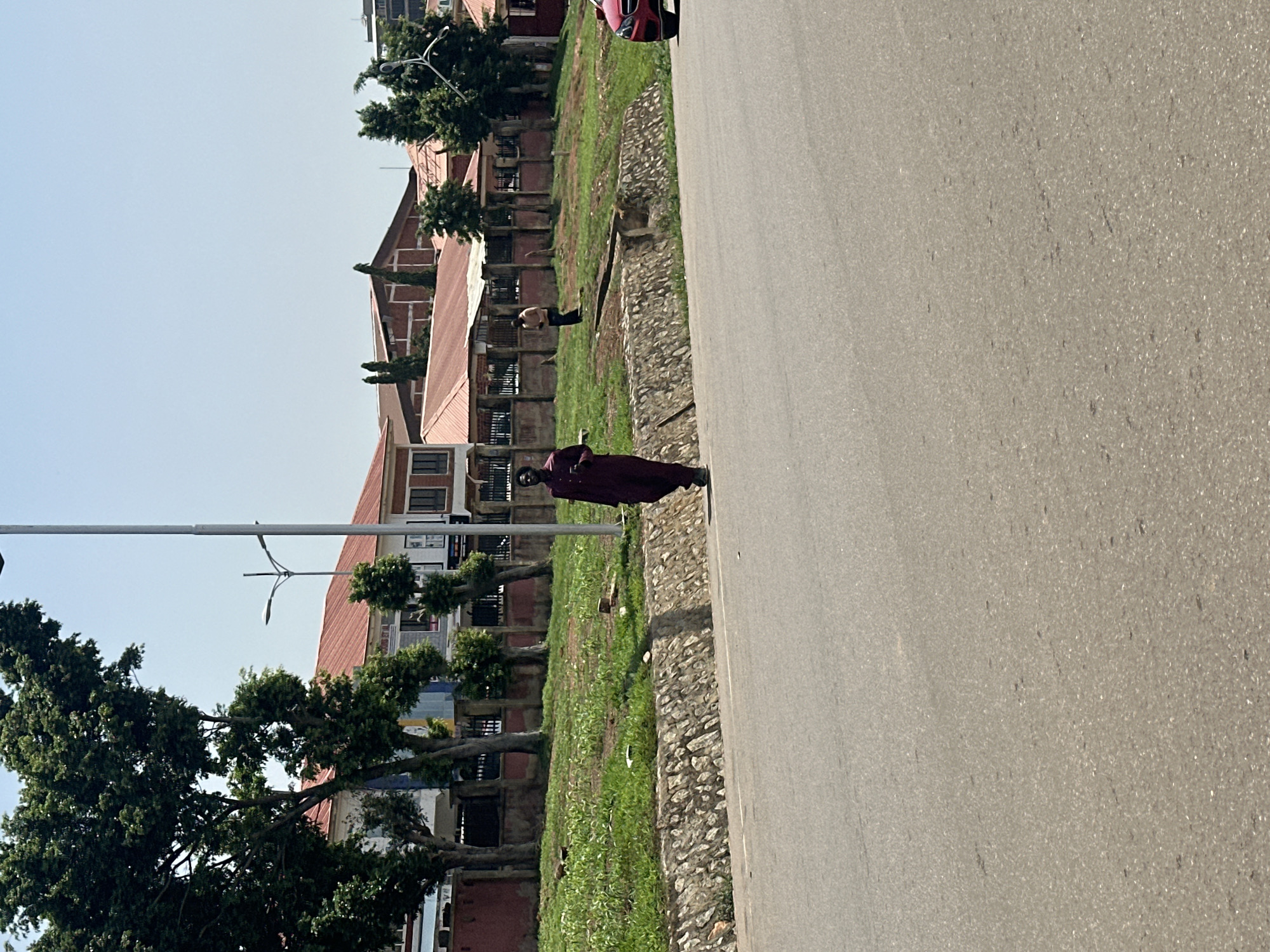 The image shows a street scene in Abuja, Nigeria. The main subjects are two people, one walking and another standing. A person in a dark maroon robe is walking on the right side of the frame, near the road. Further down the road, a person with their torso bare is standing in a grassy area. In the background, there are several buildings with red tiled roofs, and mature trees lining a walkway. A lamppost is visible in the middle ground. The ground beside the road is covered in grass and stones. The sky is clear and bright, suggesting it is daytime, likely a sunny day. The scene appears to be a public area, possibly a park or a roadside with a pedestrian path and adjacent buildings. No text is visible in the image.