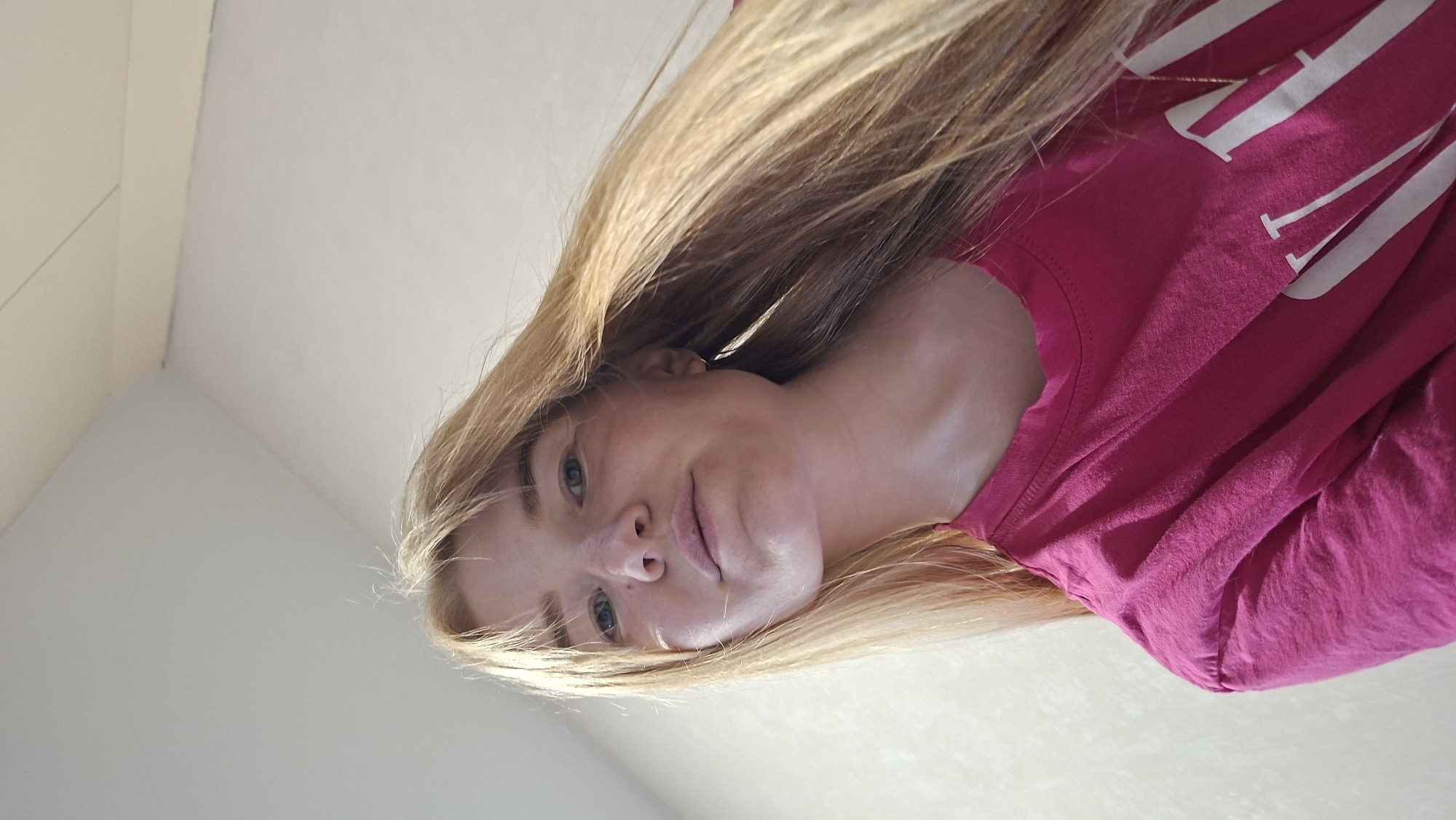 The image is a close-up, low-angle, portrait of a young woman with long blonde hair. She is wearing a bright pink t-shirt with white lettering that appears to be a logo. The woman is looking directly at the camera with a gentle, slightly tilted head and a faint smile. Her eyes are light blue, and her skin has a light complexion with some freckles visible on her nose and cheeks. The lighting suggests it is daytime, and the bright, even illumination hints at an indoor setting with good lighting, or possibly an outdoor location on a bright, overcast day. The background is a plain, light-colored surface, possibly a wall or ceiling, contributing to a minimalist aesthetic that focuses attention on the subject. There are no clear indications of the specific location within Chaikovsky, Russia, but the overall presentation is casual and personal, suggesting a candid or posed personal photograph.