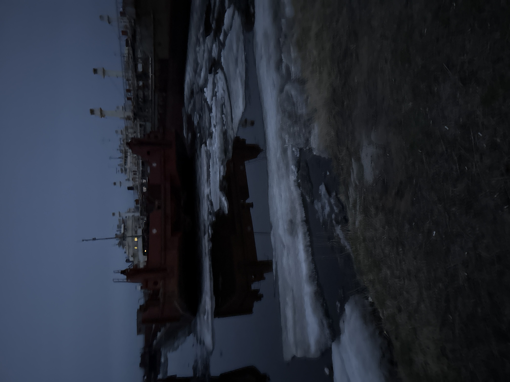 The image captures a scene at Zhatay, Russia. A large ship is visible on the left side of the frame, with its structure reflected in the water below. The sky is overcast. On the right side of the image is a grassy bank covered with patches of snow. The overall lighting suggests it is dusk or dawn.
