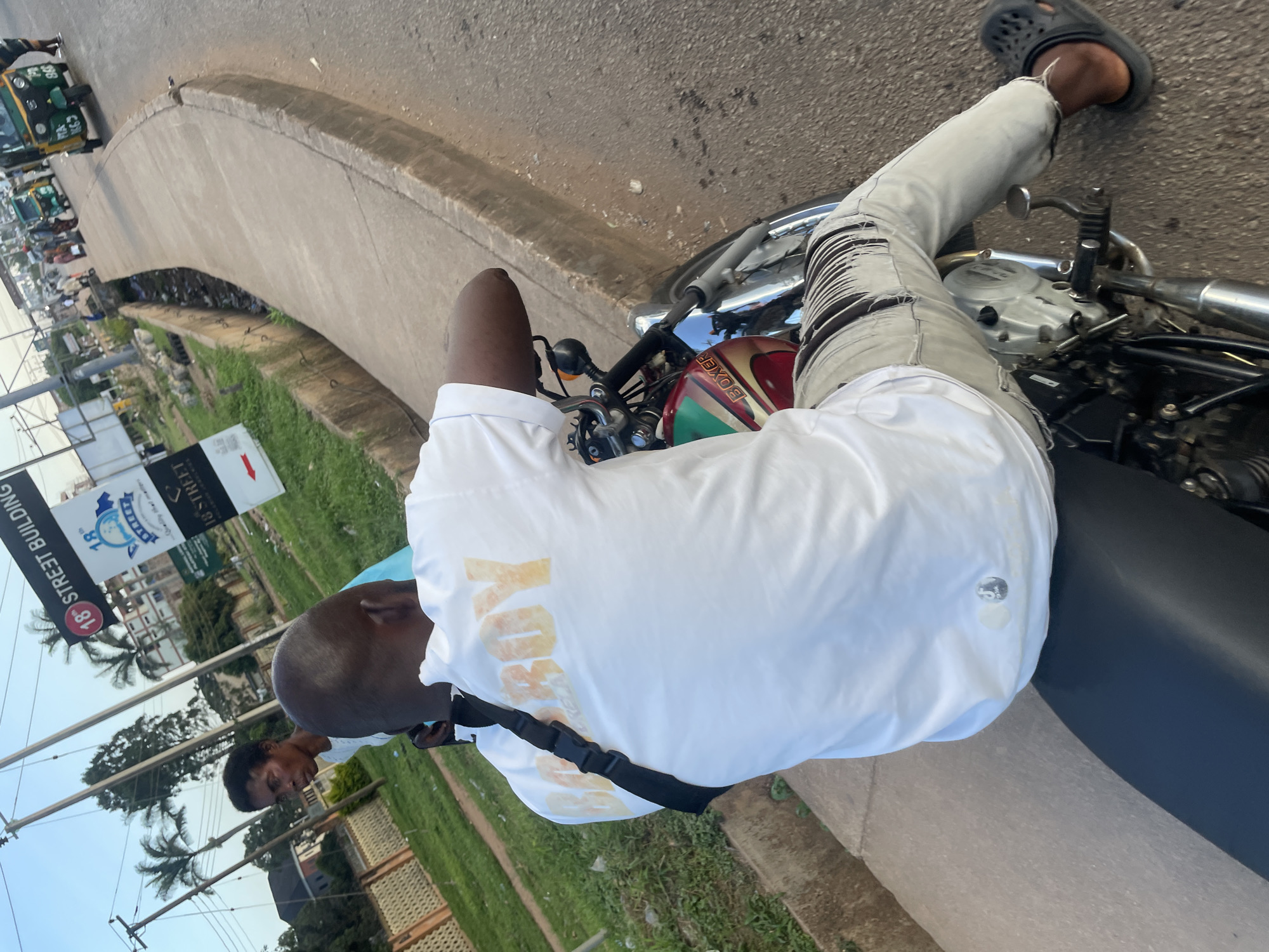 An over-the-shoulder medium shot captures a man riding a motorcycle, his back to the camera, in what appears to be a street scene in Jos, Nigeria. The man is wearing a white t-shirt with orange lettering that reads "BADBOY" and light-colored, distressed jeans. He is also wearing black Crocs on his right foot. The motorcycle's fuel tank is visible, featuring a red, green, and gold design with the word "BOXER" in gold lettering. To the left of the motorcycle, a woman in a light blue top stands on a paved walkway, looking towards the right. In the background, palm trees and buildings are visible under a clear sky. A street sign with "18th STREET BUILDING" written on it stands prominently. Several motorized tricycles, a common mode of transport in Jos, are seen on the road ahead, along with other motorcycles and pedestrians. A drainage ditch runs along the side of the road. The lighting suggests it is late afternoon or early evening.
