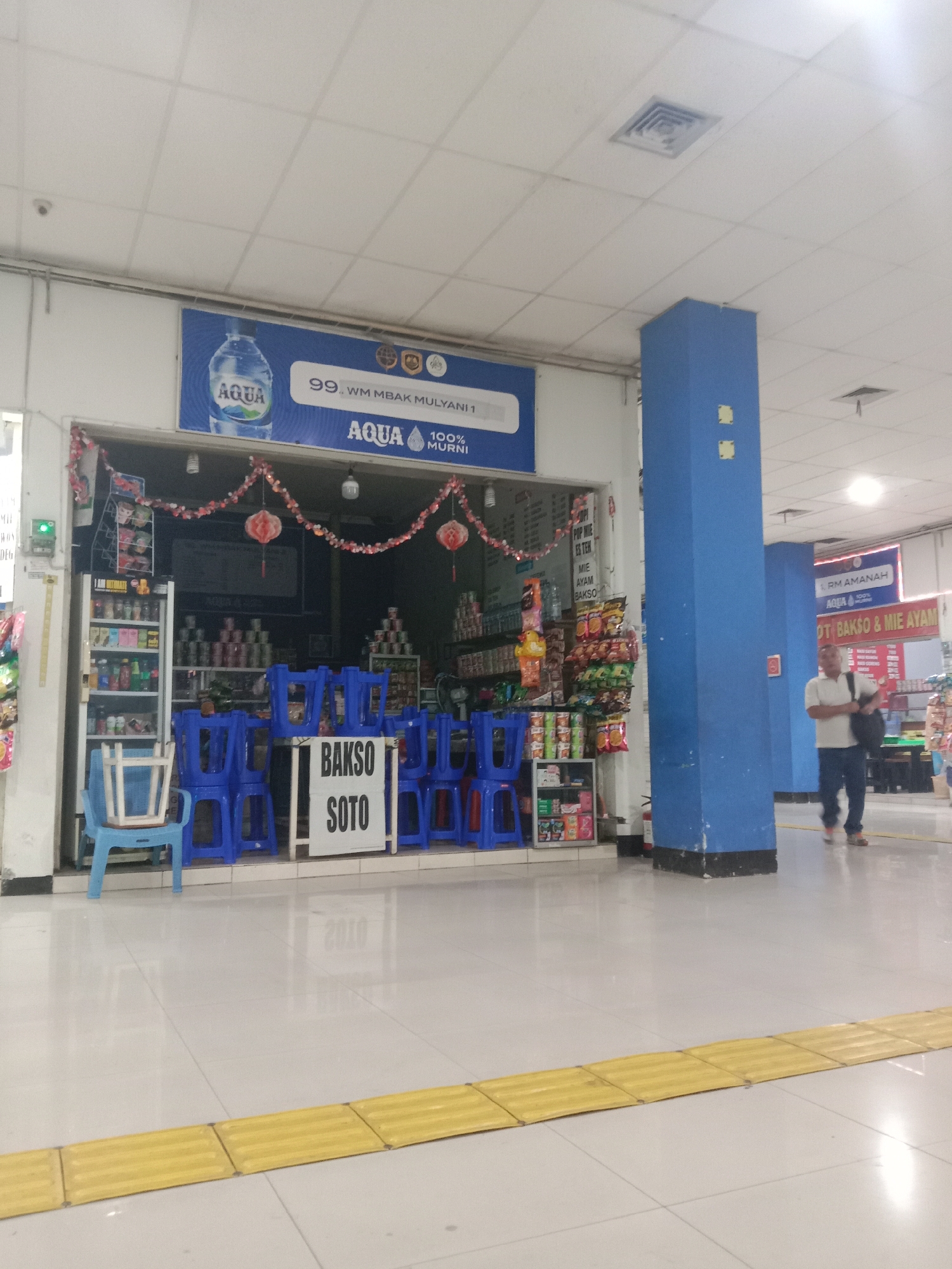 The image shows the exterior of a small food stall. The stall is open with blue plastic chairs neatly stacked inside and outside. A sign above the stall reads "AQUA" and features a bottle of water. Another sign on the stall reads "BAKSO SOTO", indicating the food items sold. Various packaged goods and drinks are displayed on shelves inside the stall. In the background, a man in a white shirt and blue jeans is walking away from the stall carrying a black bag. The floor is tiled, and a yellow tactile paving strip is visible in the foreground. The environment appears to be an indoor market or public space.