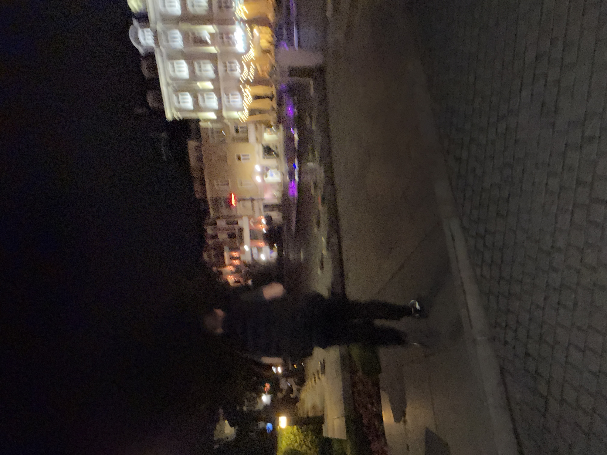 This media file is a dark, blurry photograph taken at night, rotated 90 degrees counter-clockwise. It depicts an urban or semi-urban scene illuminated by artificial lights.
The primary subject is a single person standing in the foreground, positioned on what appears to be a paved sidewalk or pedestrian area. The individual is dressed in dark clothing, and their face and specific features are obscured by darkness and blur.
In the background, a multi-story building stands prominently, with many of its windows glowing from interior lights. Decorative string lights are visible on an outdoor section of this building, possibly a balcony or terrace. Various other artificial light sources, including streetlights or lampposts, cast warm yellow, purple, and blue light across the scene. A faint red sign can be seen on one of the structures, but any text on it is illegible. Dark vegetation, such as trees or bushes, is also visible, particularly on the left side of the image. The ground in the foreground and midground is a paved surface, possibly composed of cobblestones or light-colored pavers.
The setting is clearly nighttime, characterized by the dominant artificial lighting against the dark sky. The atmosphere suggests a quiet evening in a built-up area, possibly a commercial district or hotel complex. No specific activity or event other than the person standing is discernible.