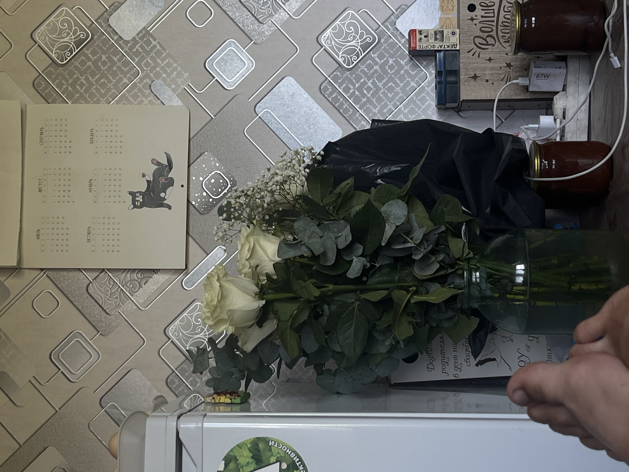 A close-up shot captures a bouquet of white roses and eucalyptus in a glass vase, set against a patterned wallpaper. To the left, a portion of a white refrigerator is visible, featuring a green circular sticker. A calendar hangs on the wall, displaying months and a cartoon cat. In the foreground, a human foot is positioned towards the lower left corner. The background reveals various objects including jars, electrical outlets, and a box with text. The overall setting appears to be an indoor domestic environment.