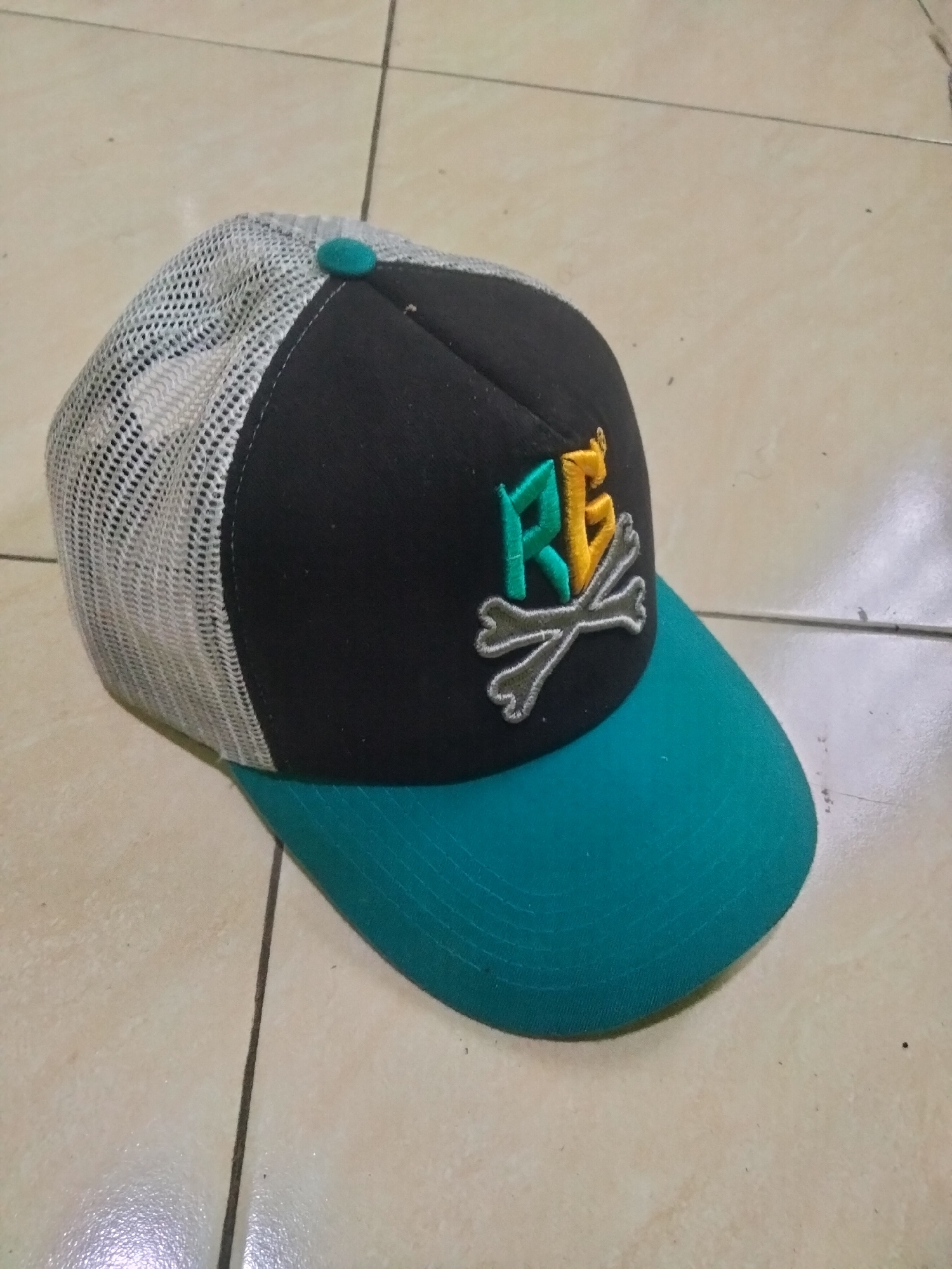 A black and teal trucker hat with a white mesh back is lying on a tiled floor. The hat features an embroidered logo with the letters "RE" in green and yellow, crossed by two grey bones. The floor is made of light-colored square tiles with dark grout lines. The scene appears to be indoors, possibly in Ketanggung, Indonesia, given the context provided. No people are visible in the image.