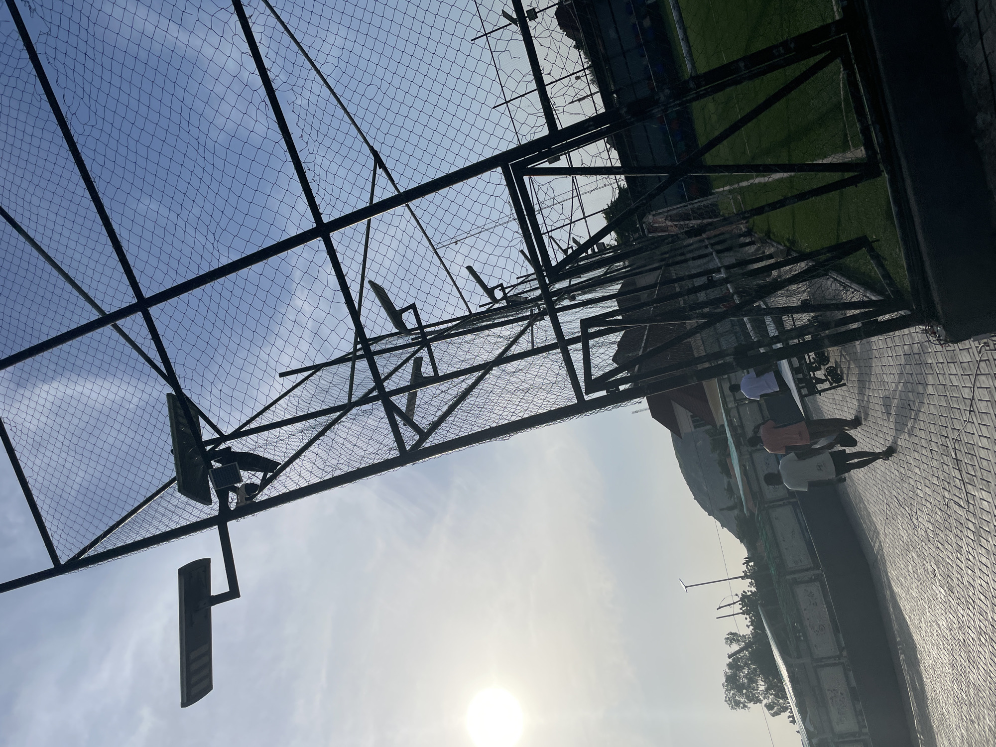 The image depicts an outdoor sports facility, likely a football pitch, located in Jalingo, Nigeria. The scene is bathed in the warm, bright light of a setting or rising sun, which casts a soft glow on the environment and creates a slight haze. The foreground is dominated by a paved walkway with a diamond pattern. To the left, a portion of a fence with metal poles and wire mesh encloses the facility. Two solar-powered streetlights are mounted on the fence structure, with one positioned prominently on the left and another further back. In the mid-ground, three individuals are walking away from the camera on the paved walkway. The person in the foreground wears a white t-shirt and black shorts. The second person is wearing an orange t-shirt, and the third person, further in the distance, is wearing a white t-shirt. A table with what appears to be a trolley or cart is visible to the right of the people. Beyond the fence, a section of a green artificial turf sports field with white markings is visible. In the background, there are buildings with reddish-brown roofs and trees, and a hill or mountain range looms in the distance under a clear blue sky with wispy clouds. The overall impression is that of a recreational area in an urban setting during daylight hours.