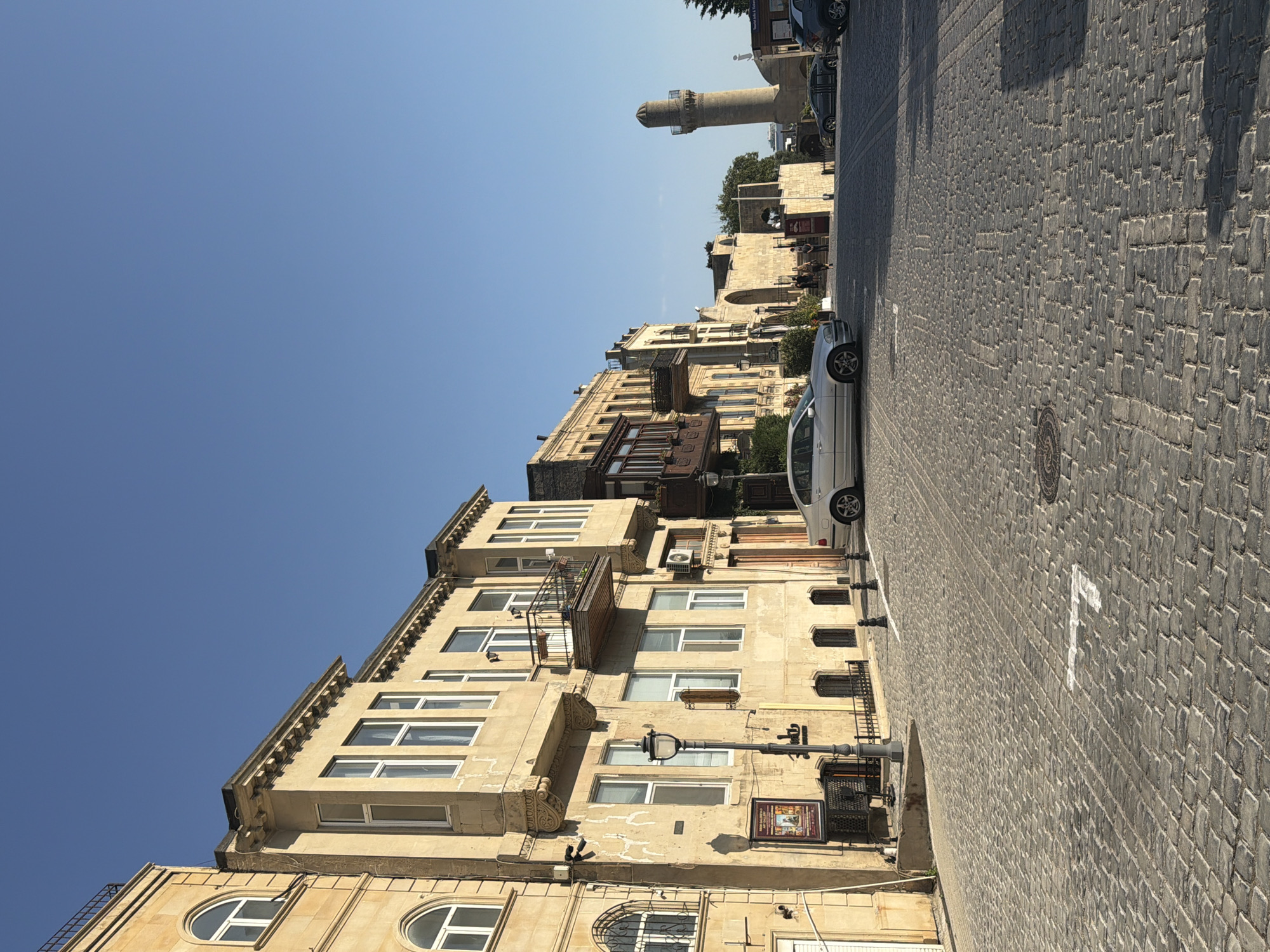 This is a ground-level, eye-level, vertical shot of a cobblestone street in the Icherisheher (Old City) of Baku, Azerbaijan, within Sabail Raion. The scene is bathed in bright, natural sunlight, casting distinct shadows. On the left, a light-colored, multi-story building with arched windows and a small balcony features classical architectural elements. A lamppost stands prominently on the sidewalk in front of this building, accompanied by a metal fence. A silver sedan is parked on the street, angled towards the right.
Further down the street, more buildings with similar architectural styles are visible, including those with wooden balconies. A tall, slender minaret, characteristic of Islamic architecture, rises in the background on the right side of the image. Several people are seen walking on the street and near an entrance to a building in the mid-ground. The street is paved with dark, worn cobblestones. The sky above is clear and blue.