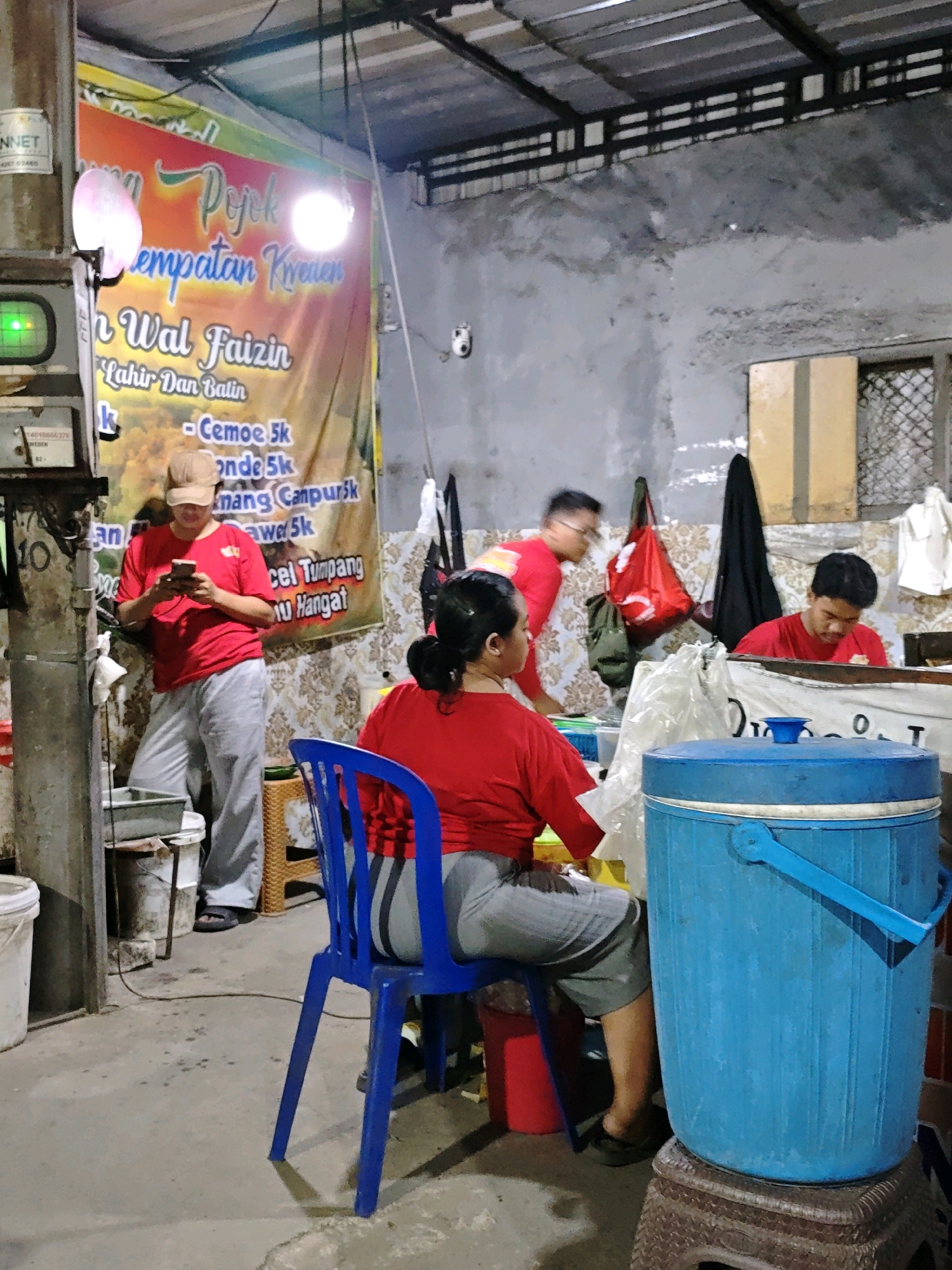 The image depicts a dimly lit food stall with a banner in the background advertising "Pojok Empatan Kreasi - Nasi Wal Faizin - Lahir Dan Batin - Cemor 5K - Ronde 5K - Nasi Tumpeng - Nasi Hangat". The stall appears to be located in Ngasem, Indonesia, indicated by the banner's text and the overall aesthetic.
In the foreground, a person wearing a red shirt and gray pants is seated on a blue plastic chair, facing away from the camera, with their legs crossed. To their right, a large blue insulated water cooler sits atop a woven stool.
Further back, a person in a red shirt and brown cap is standing and looking at a mobile phone. They are leaning against a concrete pillar with a grey bucket and other containers nearby. Another person in a red shirt is seen in the mid-ground, their face blurred and slightly out of focus, appearing to be behind a counter. In the background, a person wearing a red shirt is also visible, with a red bag hanging on a hook behind them.
The environment is rustic, with a corrugated metal ceiling supported by metal beams and rough concrete walls. The floor is also concrete. The lighting is functional, with a bright lamp illuminating the banner and general ambient light casting shadows. A green digital display is visible on the left side of the image. The overall impression is of a casual, open-air food establishment.