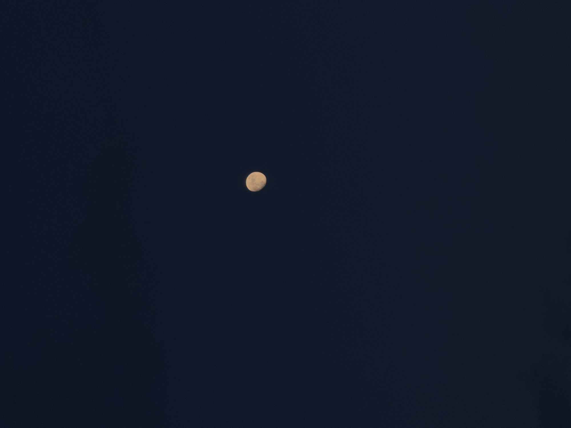 The image displays a clear night sky over Chaikovsky, Russia. The primary subject is a crescent moon, positioned slightly above the center of the frame. The moon appears yellowish-white and shows some subtle surface texture, suggesting craters or maria, though it is not sharply in focus. The sky is a deep, uniform dark blue, indicating a clear atmosphere free of clouds. There are no other visible celestial bodies like stars or planets, nor any artificial lights or identifiable land features. The overall scene conveys a serene and expansive nocturnal view with the moon as the sole focal point. There are no people, visible text, or specific activities depicted.