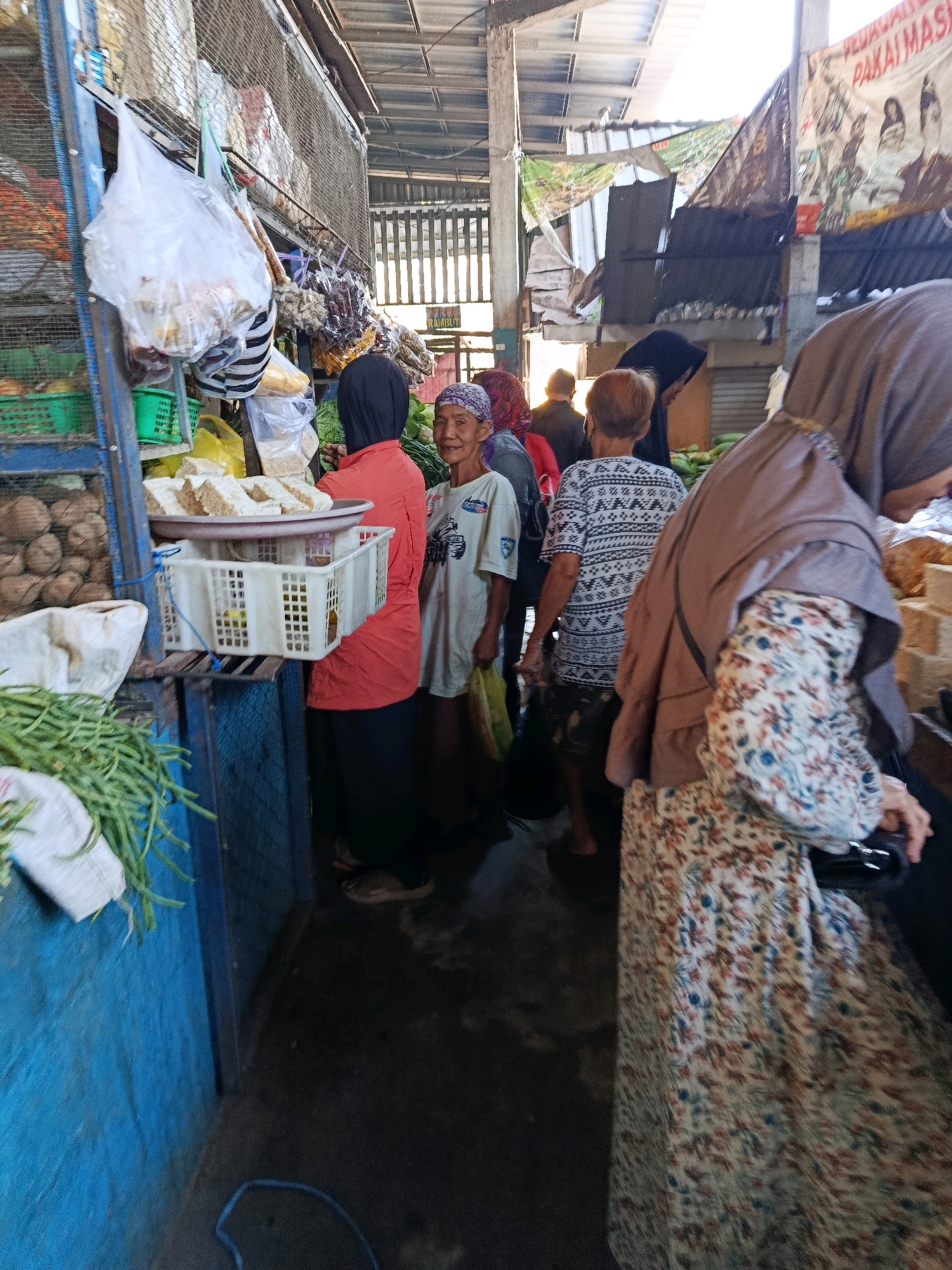 The image depicts a bustling scene within a market in Bululawang, Indonesia. The market appears to be an open-air or semi-covered structure with a corrugated metal roof. Several individuals are present, engaged in various activities typically found in a marketplace.
In the foreground, a person wearing a floral patterned dress and a brown hijab is partially visible from the side, reaching towards something on a market stall. To the left, a person in an orange shirt and black head covering is seen from behind, facing towards the right side of the frame. Beside them, an older woman with a headscarf is looking towards the camera. Another individual, wearing a light-colored t-shirt with a graphic, stands between the woman in orange and the older woman. Further in the background, other market-goers and stalls can be observed.
The market stalls display a variety of goods. In the left portion of the image, sacks of produce, possibly potatoes, are visible in green baskets. Another stall has stacked tempeh, a fermented soybean product, on a tray and in white plastic crates. Bags of dried goods and what appear to be spices or herbs hang from the ceiling or are displayed on shelves. The ground appears to be concrete and is somewhat damp, suggesting it may be a humid environment or has recently been cleaned.
The overall atmosphere is one of a vibrant, working market with everyday commerce taking place. The lighting suggests it is daytime, with sunlight filtering in from the open areas of the market. The presence of signs in Indonesian language further confirms the location.