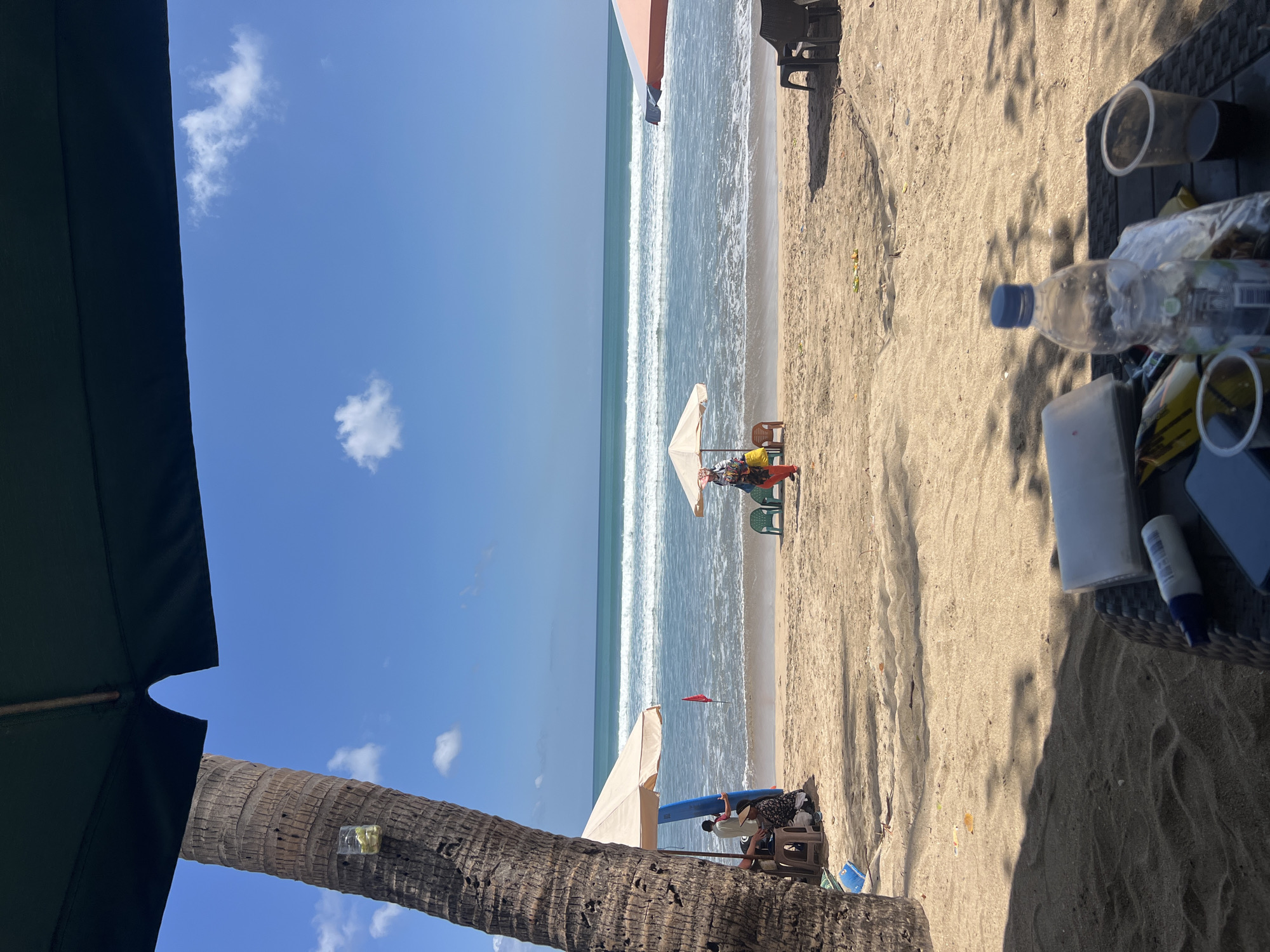 This is a medium shot, eye-level, outdoor photograph of Kuta Beach in Kuta, Indonesia, on a sunny day. The foreground is dominated by a table with several items, including a plastic bottle of water, two disposable cups (one with a dark liquid), a package of something, and a white tissue box. In the midground, there is a sandy beach with a few beach umbrellas, chairs, and people. A person in orange pants and a colorful top stands near a light-colored umbrella, holding a bag. To the left, under a tan umbrella, a person sits on a chair next to a blue surfboard. A red flag is planted in the sand. The background features the ocean with gentle waves and a clear blue sky with a few white clouds. A large palm tree is visible on the left side of the frame, with a glass containing a yellow substance attached to its trunk. The overall impression is a relaxed beach scene.