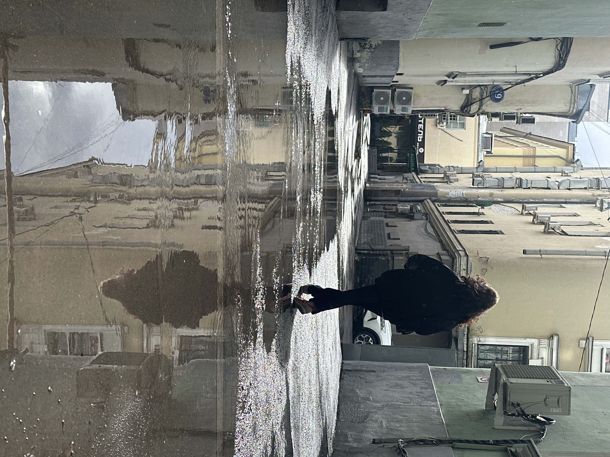 A person with dark, curly hair walks away from the camera down a narrow, wet alleyway in Moscow, Russia. The person is wearing a black jacket and dark pants. The alleyway is lined with multi-story buildings, some of which have air conditioning units mounted on their exteriors. Puddles of water on the ground reflect the buildings and the sky. A white car is parked in the distance. The scene appears to be in a historic part of the city.
