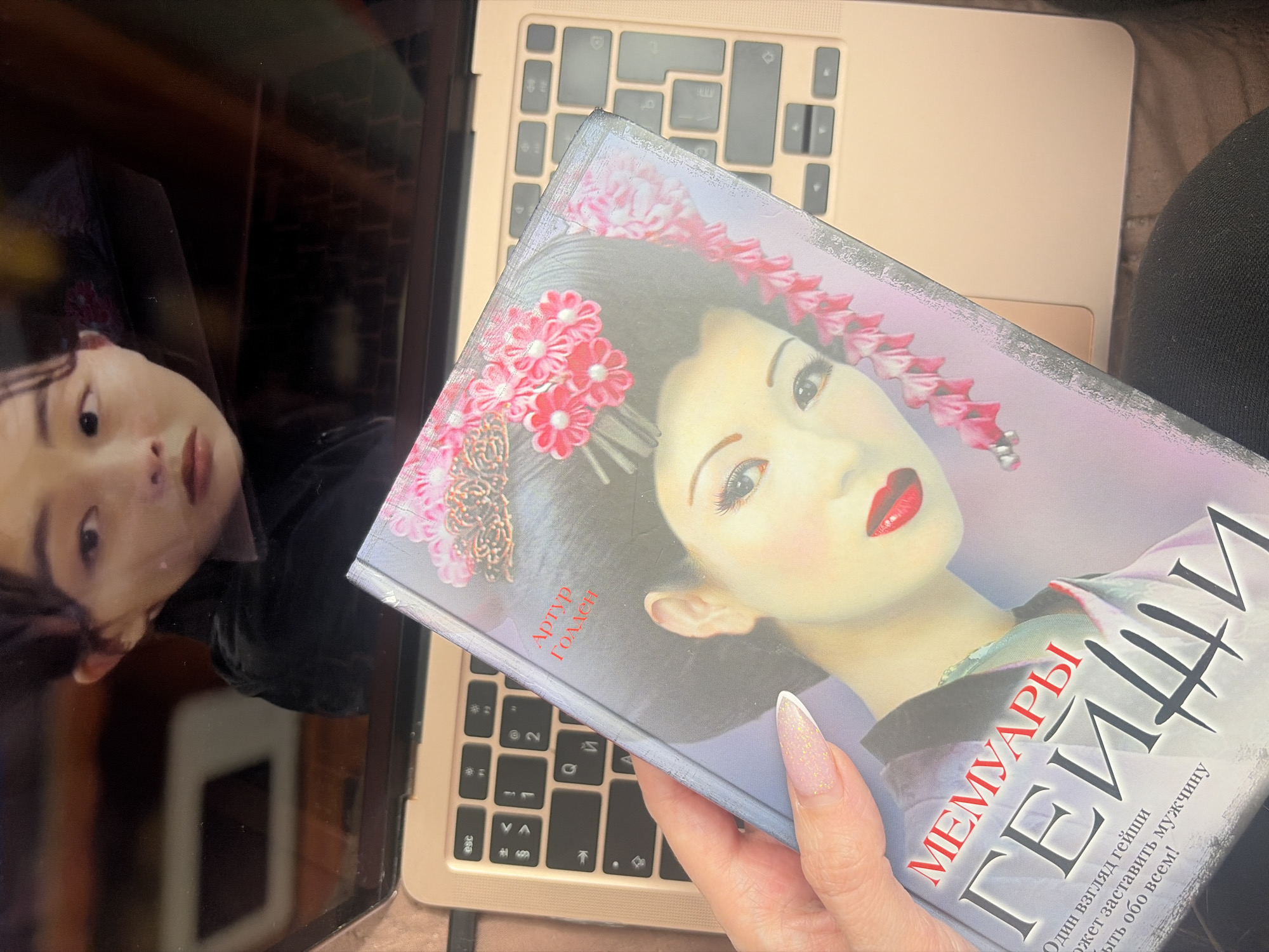 The image presents an overhead shot focusing on a person's hand holding a book over a laptop, likely in Saint Petersburg, Russia. The scene captures a moment of reading or contemplation.
The main subjects include a closed, light gold-colored laptop, whose dark screen reflects the upside-down face of a young person with dark hair and an earnest expression. The reflection is quite clear, showing the person's eyes, nose, and mouth. Resting on the laptop, a left hand with manicured nails (featuring a sparkling pink and white French tip design) holds a hardcover book.
The book's cover is a prominent visual element, displaying a stylized illustration of a geisha with pale skin, vivid red lips, and an elaborate traditional hairstyle adorned with pink floral kanzashi hair ornaments. The title "МЕМУАРЫ ГЕЙШИ" (Memoirs of a Geisha) and the author "Артур Голден" (Arthur Golden) are clearly legible in Cyrillic script. A partial tagline below reads: "Один взгляд гейши / может заставить мужчину / забыть обо всем!" (A geisha's glance / can make a man / forget everything!).
The laptop keyboard, visible underneath the book, also displays Cyrillic characters (e.g., "Й", "А", "V", "З", "esc"), reinforcing the Russian context and aligning with the provided location of Saint Petersburg. The overall setting appears to be indoors, under ambient lighting, suggesting a personal and relaxed environment.