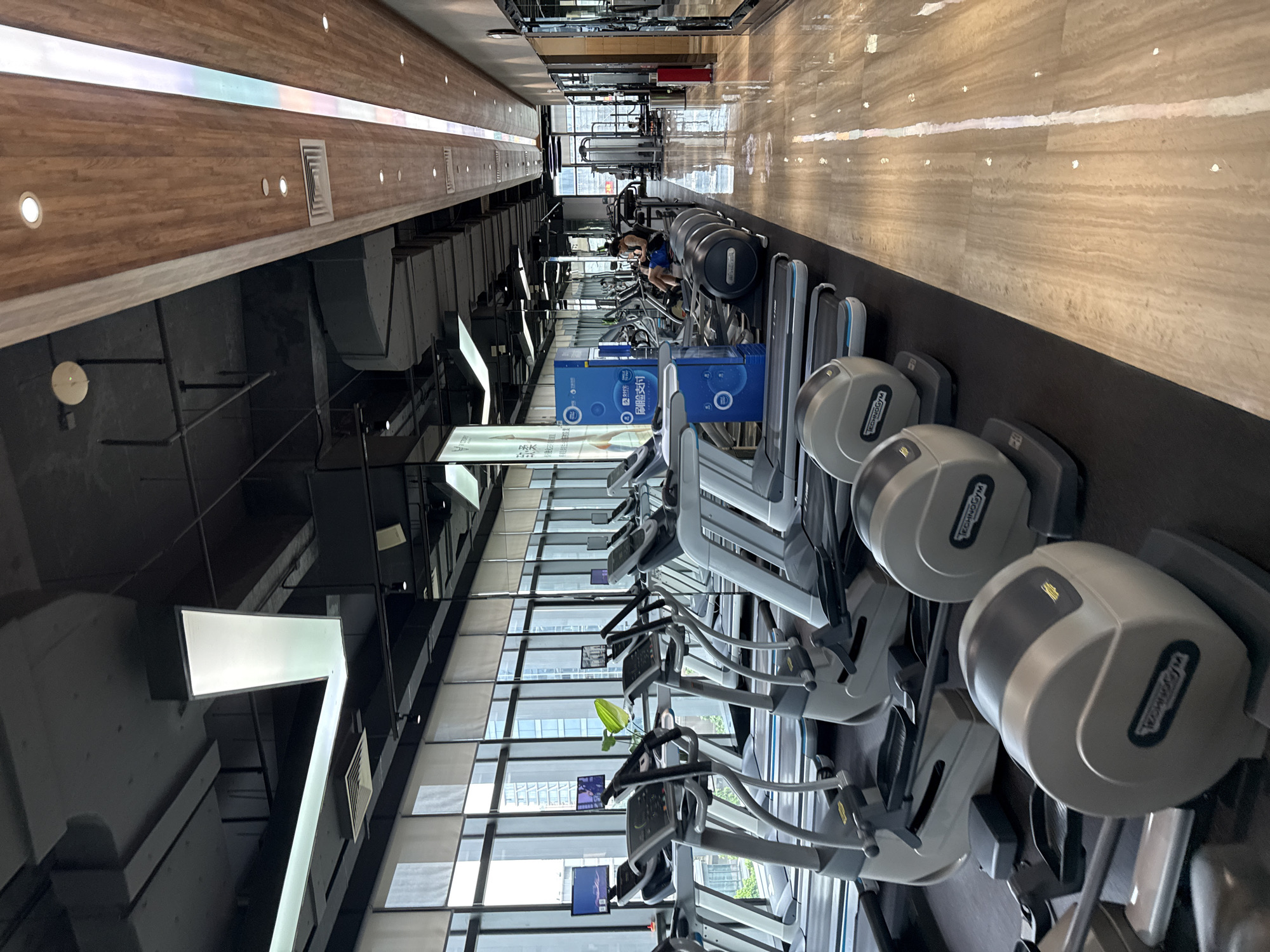 A gym with rows of cardio equipment, including treadmills and ellipticals, is captured from a slightly elevated perspective looking down a long hallway. The room is well-lit with artificial lighting. Large windows line one side of the gym, revealing an urban cityscape with modern buildings. The floor is a polished, light-colored marble, reflecting the lights and equipment. Several pieces of exercise machinery are visible, with a few being occupied by gym-goers. In the background, some weightlifting equipment and mirrors are present. The overall impression is of a modern, clean, and well-maintained fitness facility.