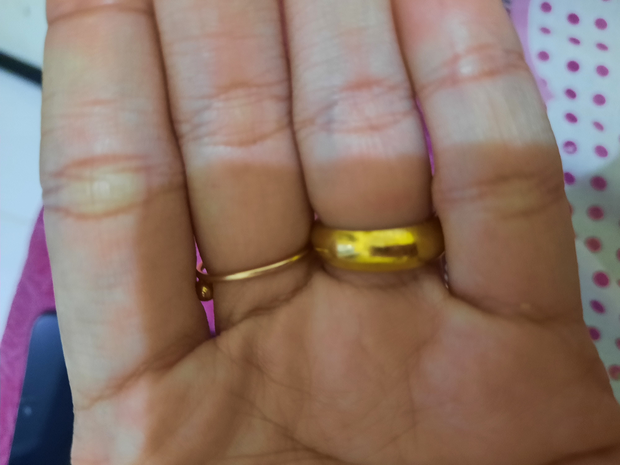 A close-up shot shows a person's hand wearing two gold rings, one on the ring finger and another on the index finger. The hand is positioned over a pink and white polka-dotted surface. The background is blurred, but appears to be a table with a dark object on it. The lighting is soft, creating a warm glow on the hand and rings. The image is focused on the hand and rings, highlighting their details and texture. The location in Lhokseumawe, Indonesia is not discernible from the provided image.