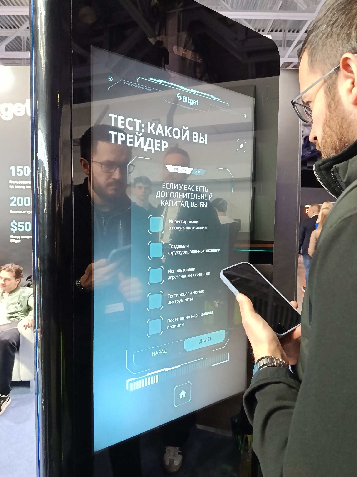 Here is a description of the image:
The image shows a man in eyeglasses interacting with a touch screen kiosk. The kiosk is black with blue accents and displays a test titled "Test: Which Trader Are You?" in Russian. The man is holding a smartphone in his left hand. The kiosk displays a multiple-choice question: "If you have additional capital, what would you do?" with options like "Invest in popular stocks", "Create structured positions", etc. The Bitget logo is visible at the top of the screen. In the background, there are other people and exhibition setups. Based on the language and context, it is likely the photo was taken at an exhibition or conference in Moscow, Russia.