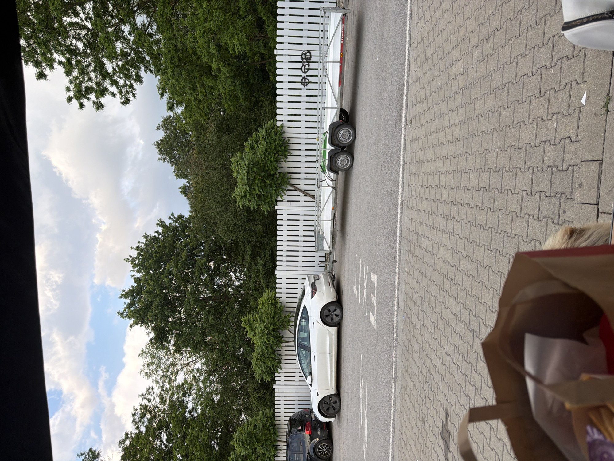 The image presents an outdoor scene in Osikovitsa, Bulgaria, captured from a low vantage point, likely from inside a vehicle. In the immediate foreground, on the left, the upper portion of a brown paper bag is visible, containing a white wrapper and a reddish item. To the right of the bag, a light-colored, possibly blond, head (suggesting a person or an animal, like a dog) is partially seen, alongside a white bag with a black zipper on the far right.
The ground beyond the immediate foreground is paved with grey, interlocking concrete pavers, leading to a section of asphalt. A white painted 'EXIT' sign, oriented for traffic departing the scene, is visible on the asphalt, accompanied by an arrow indicating a left and forward turn.
In the midground, two vehicles are parked in tandem, facing away from the viewer. On the left, the rear of a dark-colored (likely black) minivan or SUV is visible, featuring a partial Bulgarian license plate ending in "ES 7780 PP". Immediately to its right, a white Tesla Model 3 is parked, also facing away, equipped with black multi-spoke wheels. A tow hitch extends from the rear of the Tesla, connecting it to a long, empty, two-axle utility trailer. The trailer is predominantly silver/white with green wheel arches over its two wheels on the visible side, and the brand "TEMAREB" is discernible on its side.
The background is dominated by a continuous white picket fence that spans horizontally across the frame. Behind the fence, a dense line of various green trees with rich foliage forms a natural backdrop. Above the trees, the sky is partly cloudy, featuring prominent white cumulus clouds interspersed with patches of blue sky, indicating daytime conditions.