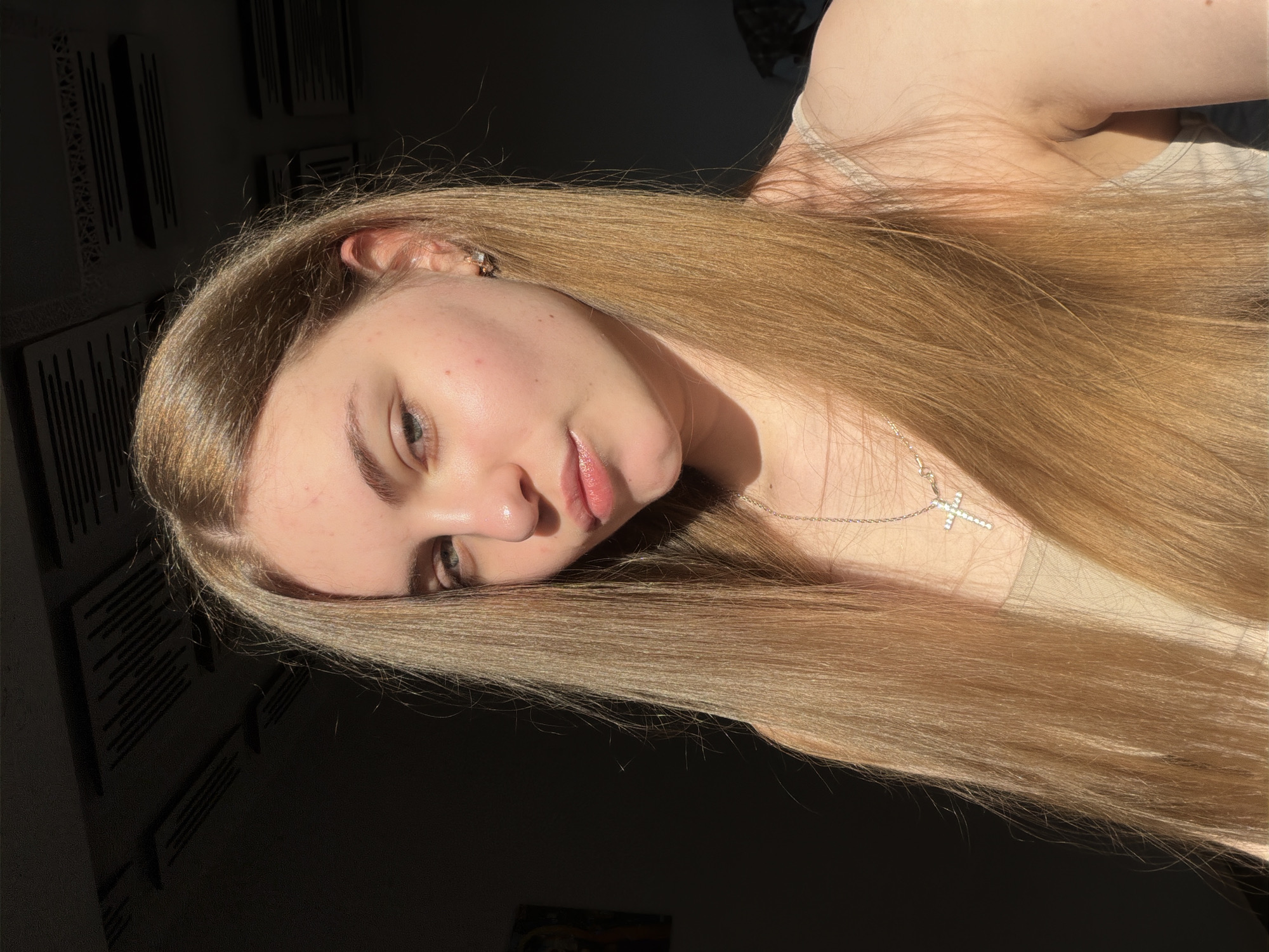The image is a close-up, chest-up selfie of a young woman with long, straight, light brown hair. She is wearing a light-colored tank top and a silver necklace with a cross pendant. She has light-colored eyes and is looking towards the camera with her lips slightly parted. The lighting suggests it is daytime, with sunlight illuminating her face and hair, creating a warm glow. The background is dark and out of focus, with some indistinct shapes that could be ceiling elements or furniture. There is no discernible location in Dnipro, Ukraine, or any other specific geographical context provided by the visual elements within the image itself.