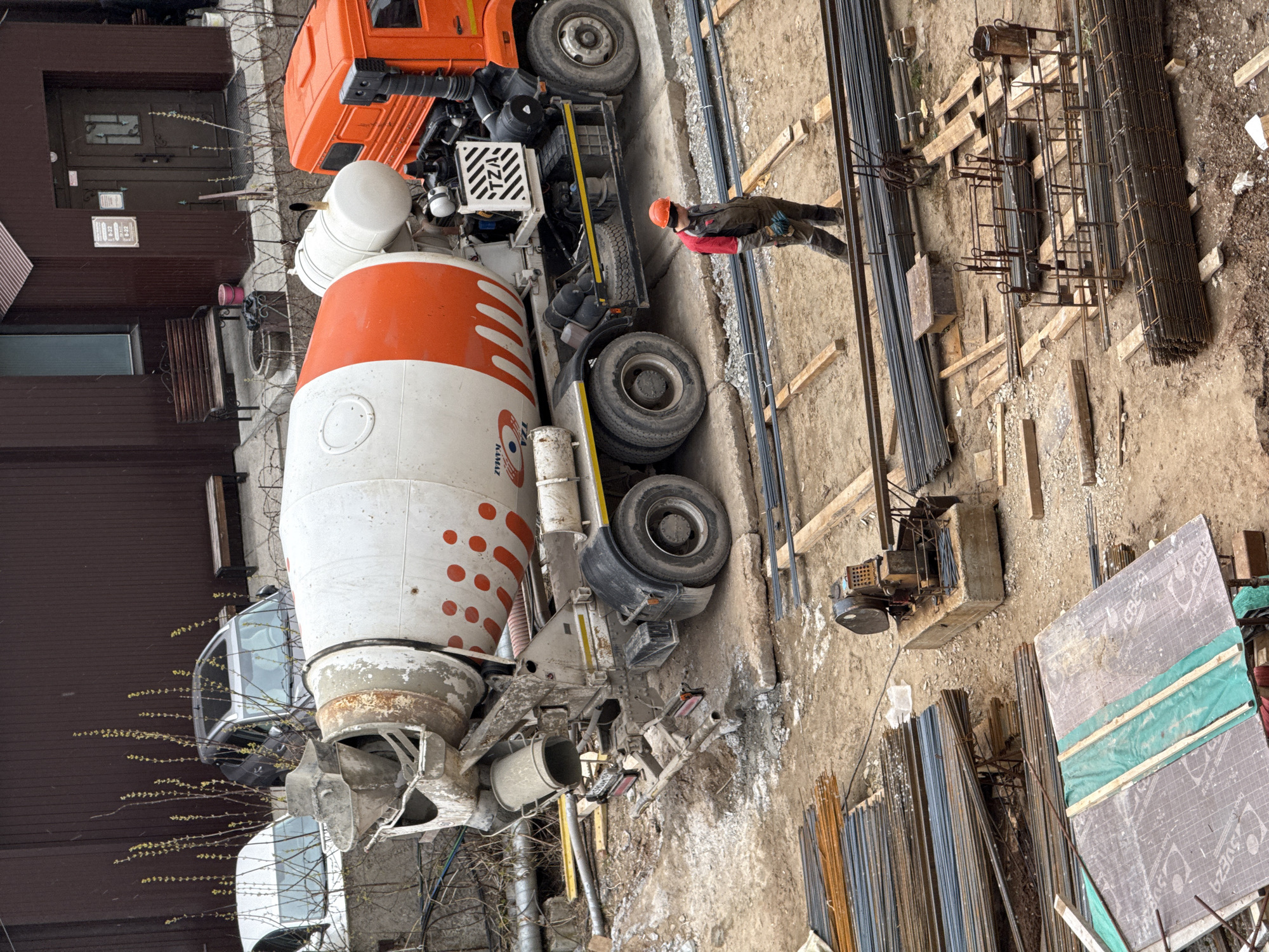 The image captures a construction site during the daytime, likely under overcast conditions, given the soft, diffused lighting. A large orange and white concrete mixer truck, bearing the text "КамАЗ" (KAMAZ) on its side, is prominent on the left side of the frame. The truck's wheels and chassis show signs of dirt and use, typical of a construction vehicle.
In the center-right, a construction worker stands on the uneven dirt ground. The worker is dressed in dark work attire, a red long-sleeved shirt or jacket, and an orange hard hat, indicating adherence to safety standards. They appear to be observing the work area or materials.
The ground is a mix of dirt and sand, scattered with various wooden planks, beams, and scaffolding elements. Extensive quantities of rebar are visible across the site; some are neatly bundled in long stacks, while others are pre-assembled into cages, indicating preparations for reinforced concrete structures such as foundations or pillars. A small piece of machinery, possibly for bending or cutting rebar, is also present on the ground.
In the background, on the far left, part of a brown building with a dark entrance and windows is visible. A small sign on the building displays "8-22" repeatedly, which likely denotes operating hours. Bare tree branches with small green buds are seen near the building, suggesting early spring. On a gray insulation board lying on the ground, the text "ЭКОДЕЗА" (EKODEZA) and "ПБС" (PBS) can be discerned.
The scene conveys an active construction environment, focused on concrete and rebar work.