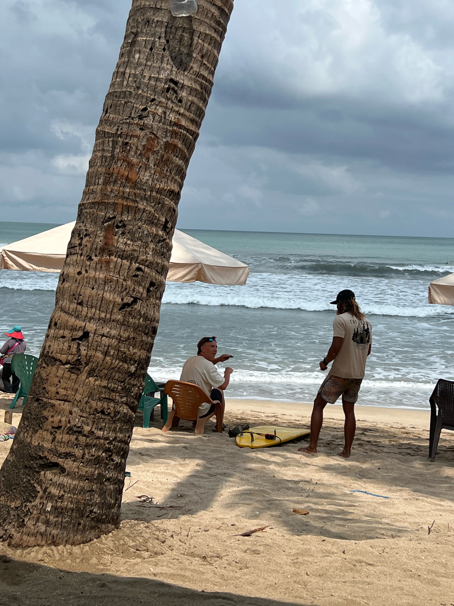 On a cloudy day at Kuta Beach, Bali, Indonesia, a thick palm tree trunk dominates the foreground. In the middle ground, two men stand on the sand near the ocean. One man, seated in a brown plastic chair, gestures with his hand as he speaks to the other man. The second man, facing away from the camera, wears a tan t-shirt with a graphic on the back, a black baseball cap, and patterned shorts. A yellow surfboard rests on the sand in front of him. Further back, a light-colored umbrella shades a seating area, and the ocean waves crash onto the shore. In the background, the overcast sky meets the sea. To the left, another umbrella is visible, and a person in a blue hat sits in a green plastic chair.