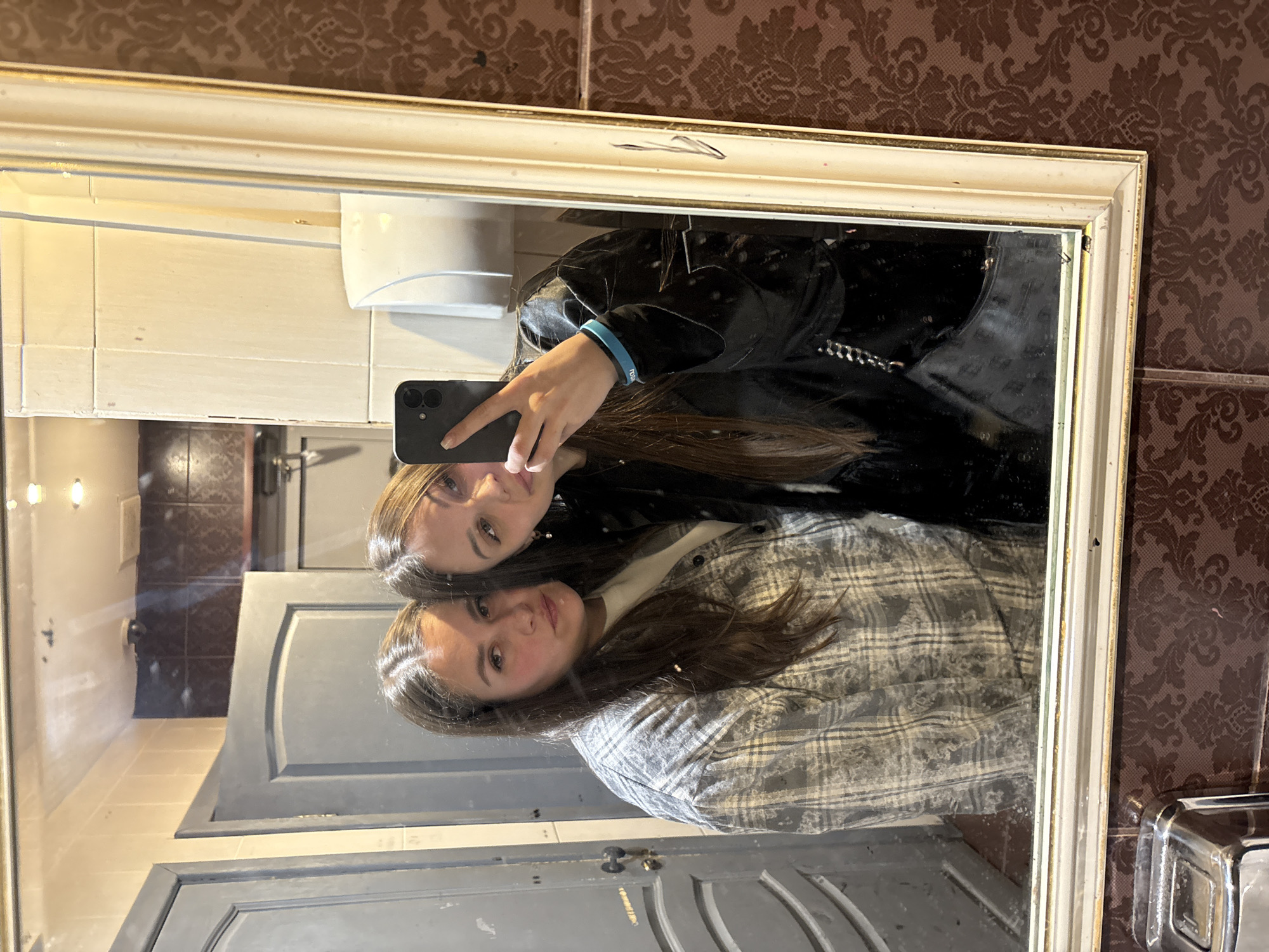 A selfie is taken by two women in a bathroom reflected in the mirror. The woman in the upper part of the mirror is wearing a black top and holding a black smartphone. The woman in the lower part of the mirror is wearing a light grey plaid shirt. The bathroom has light brown textured wallpaper, white walls, white countertops, and grey doors. The image was taken in a bathroom in Izhevsk, Russia.